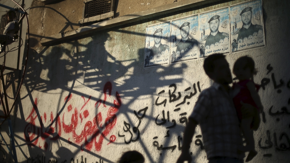 A Palestinian man carries his son as the shadow of Palestinian children playing on a swing is cast against a wall in the east of Gaza City
