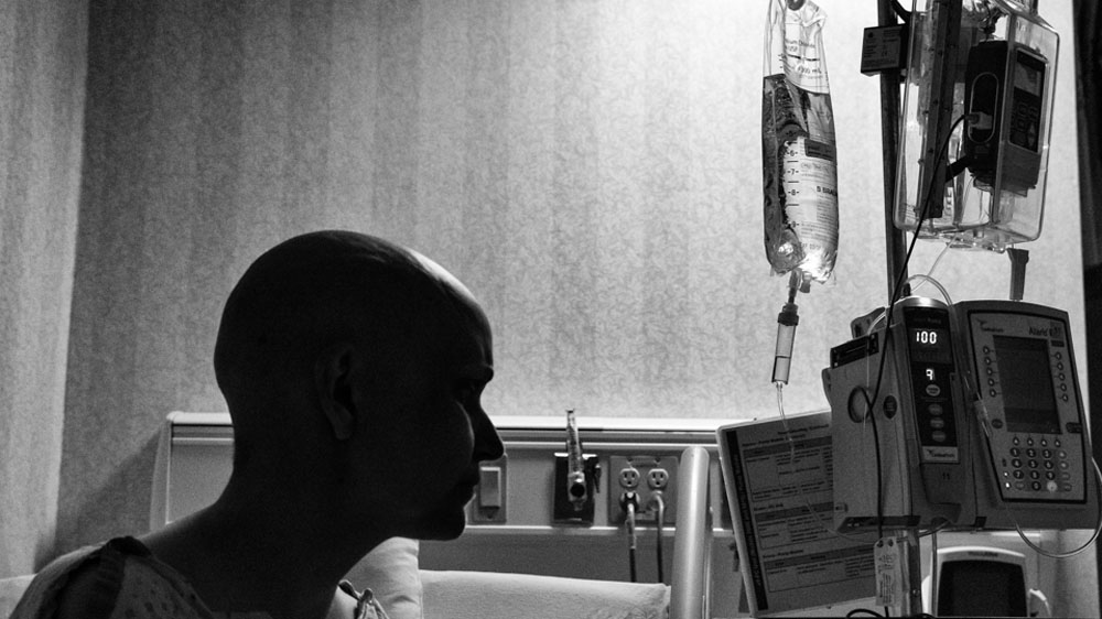 Jennifer sits in her dark hospital room, connected to machines that deliver chemotherapy, pain medication, and other fluids into her body. [Angelo Merendino/Al Jazeera] 