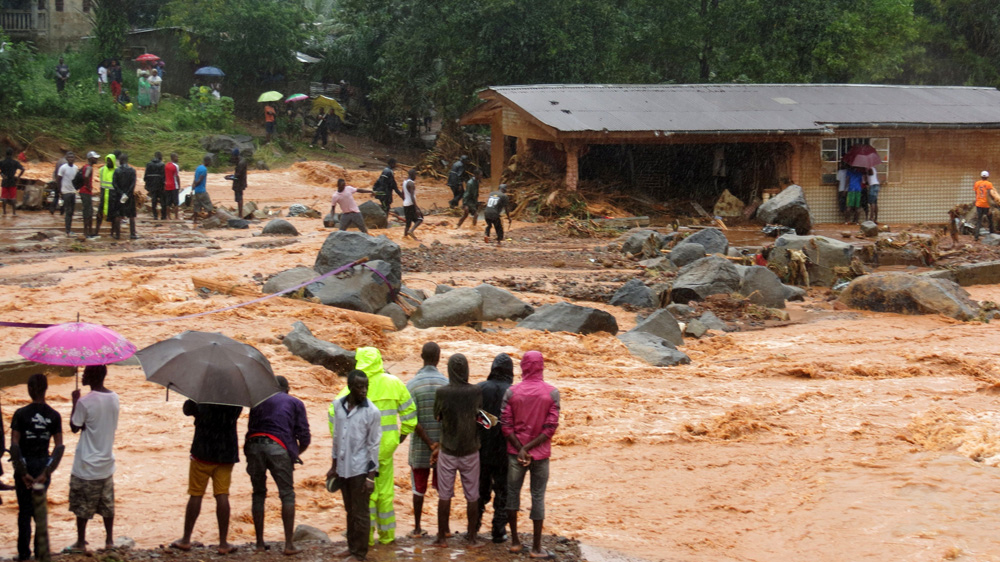 Bystanders look on as floodwaters rage past a damaged building in an area of Freetown on August 14, 2017 [Mohamed Saidu Bah/AFP/Getty Images]
