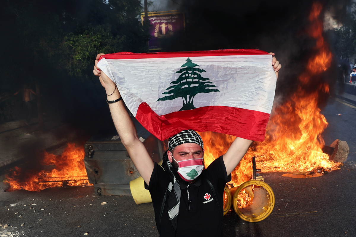 An anti-government protester holds up a Lebanese flag as others burn tires and garbage containers to block a main road, during ongoing protests against the Lebanese government, in Beirut, Lebanon, Sat