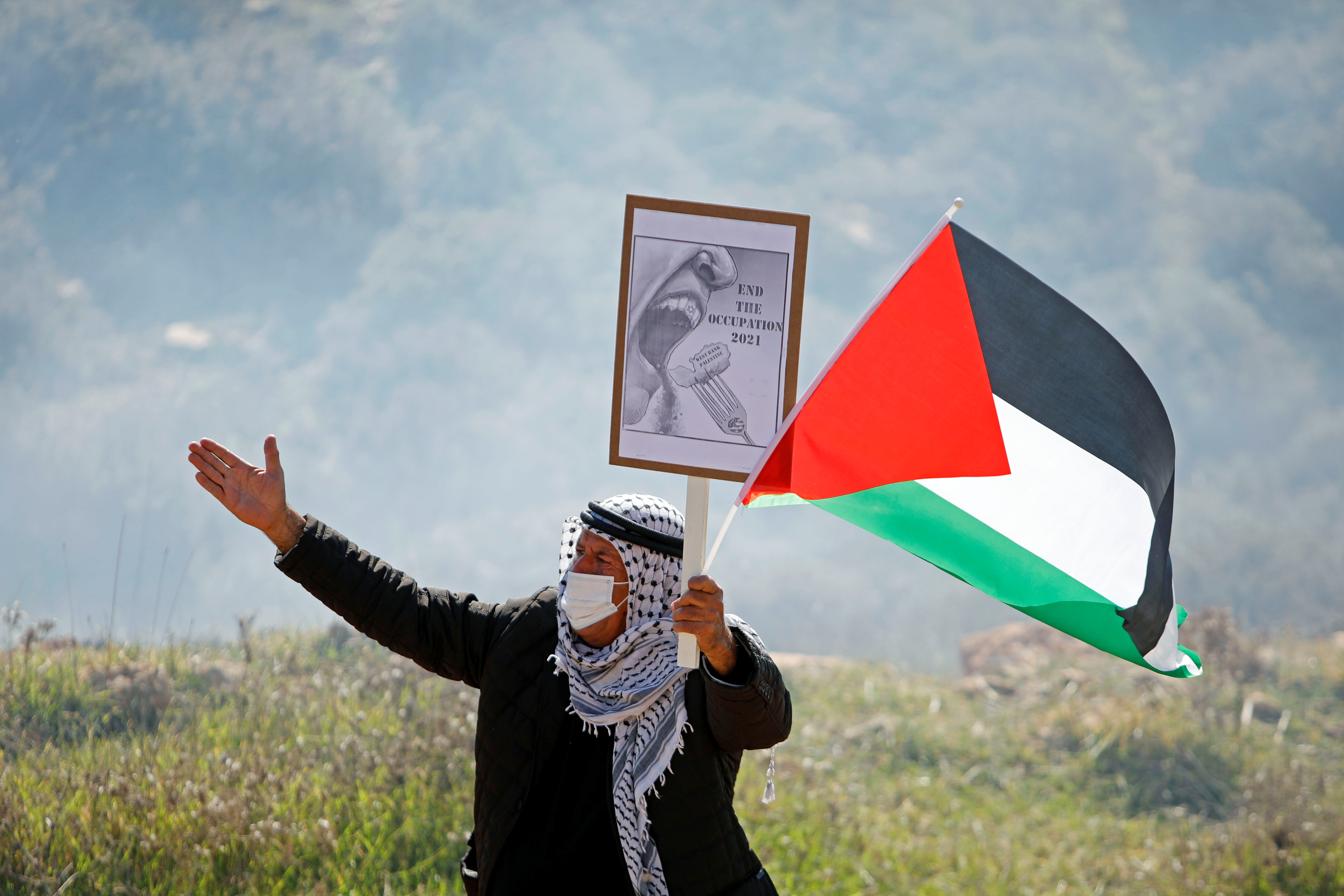 A demonstrator wearing a mask holds a banner and a Palestinian flag during a protest against Israeli settlements, in Beit Dajan, in the Israeli-occupied West Bank on January 8, 2021 [Raneen Sawafta/Reuters]