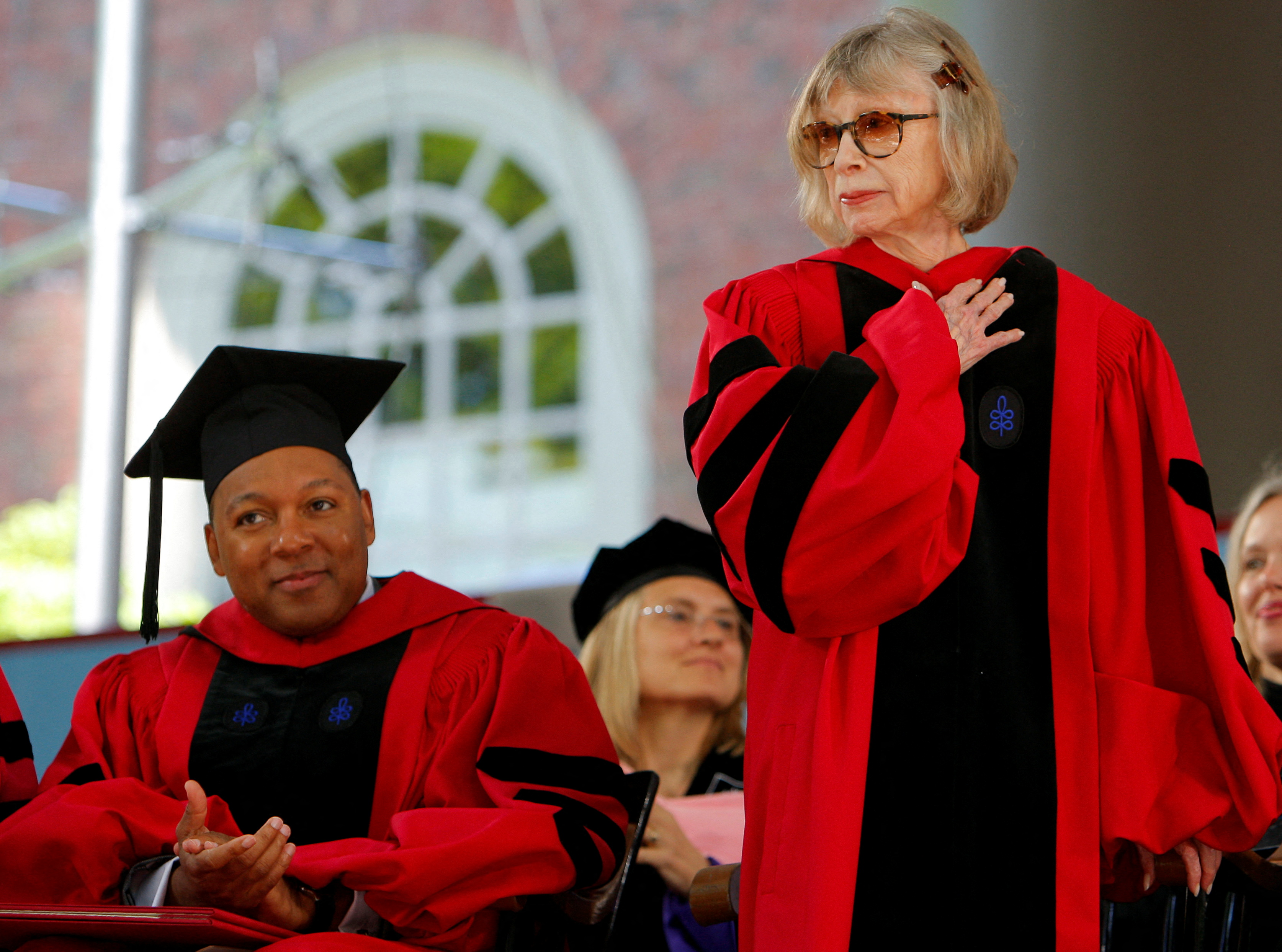 Wynton Marsalis, seated in cap and gown, applauds Joan Didion, robed and standing, on a stage.