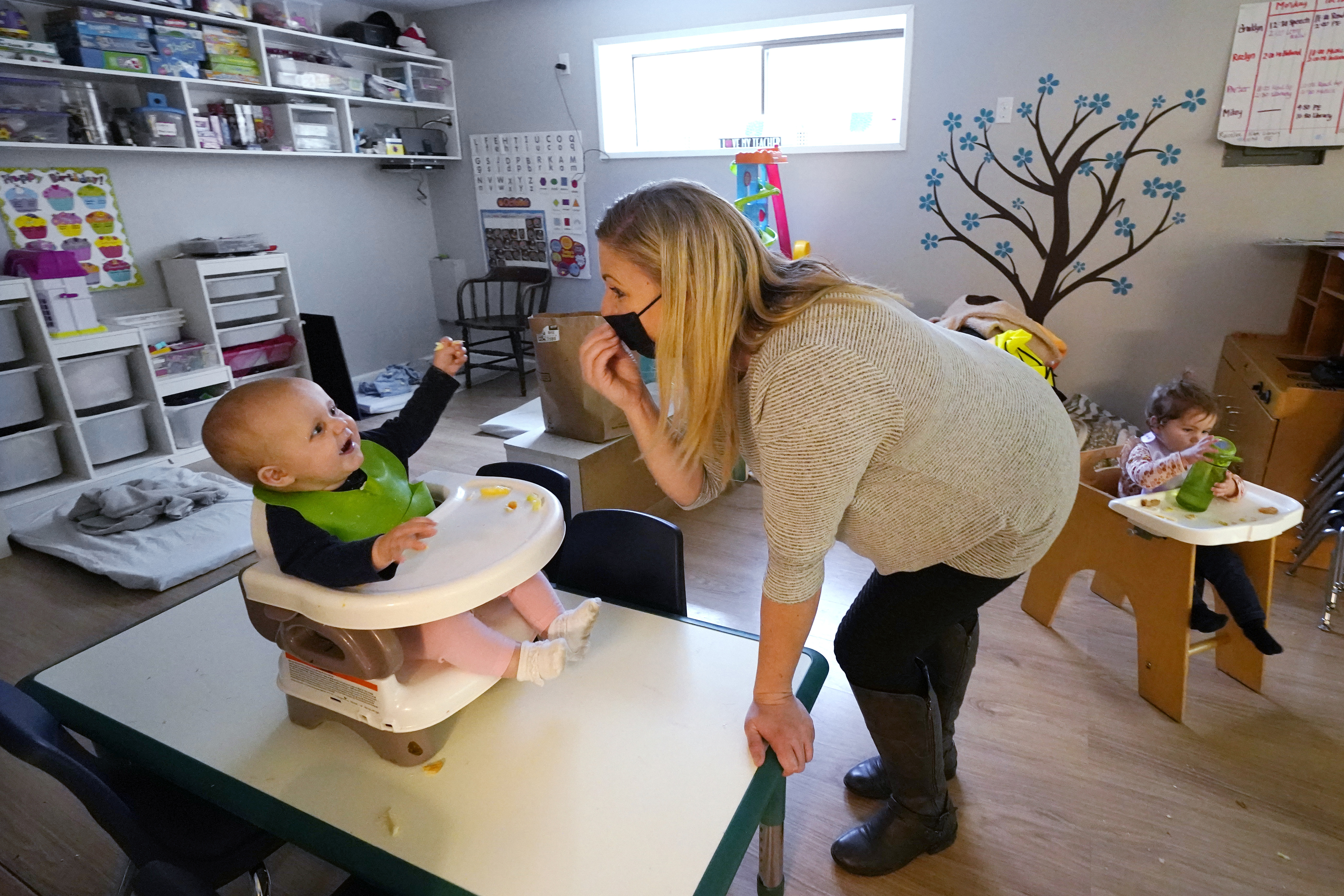 Amy McCoy signs to a baby about food as a toddler finishes lunch behind at her Forever Young Daycare facility, Monday, Oct. 25, 2021, in Mountlake Terrace, Wash. Child care centers once operated under the promise that it would always be there when parents have to work.