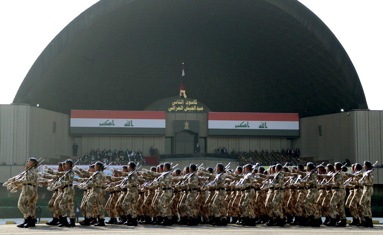 Iraqi armed forces are seen marching in a ceremony outside with two large Iraqi flags displayed above them