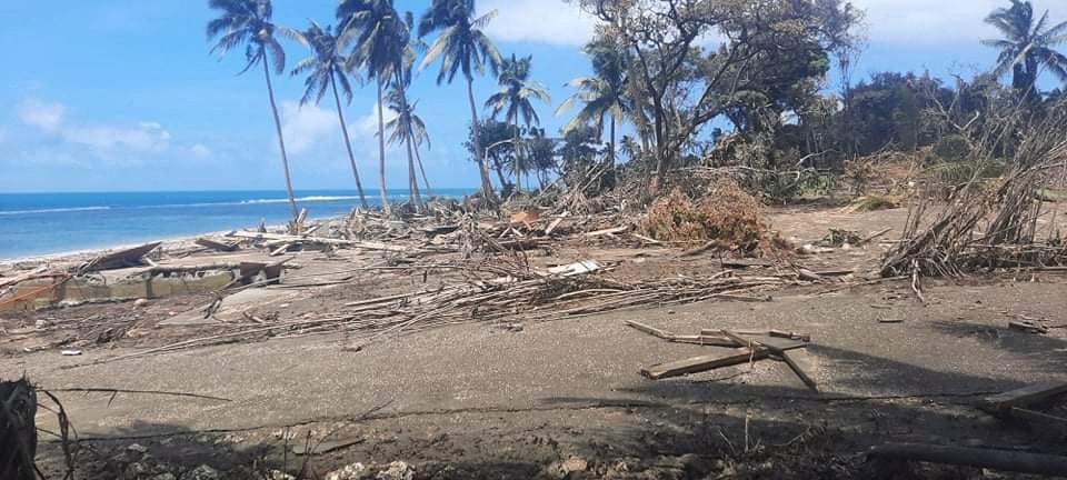 A beach in Nuku'alofa, with palm trees behind and a blue sky, covered with volcanic ash and debris from the tsunami