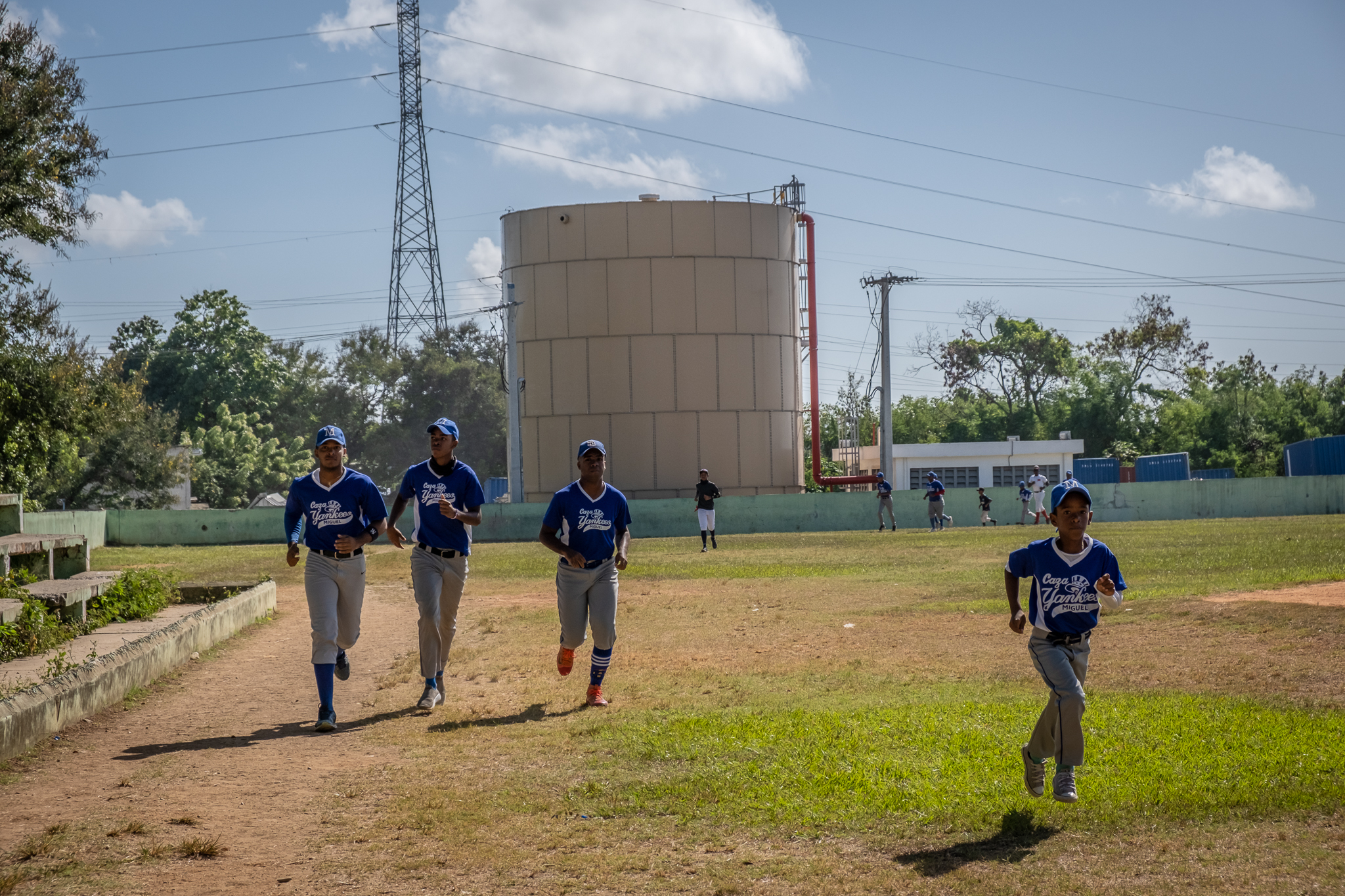 Boys playing baseball jog on a pitch that lies beside chemical storage containers