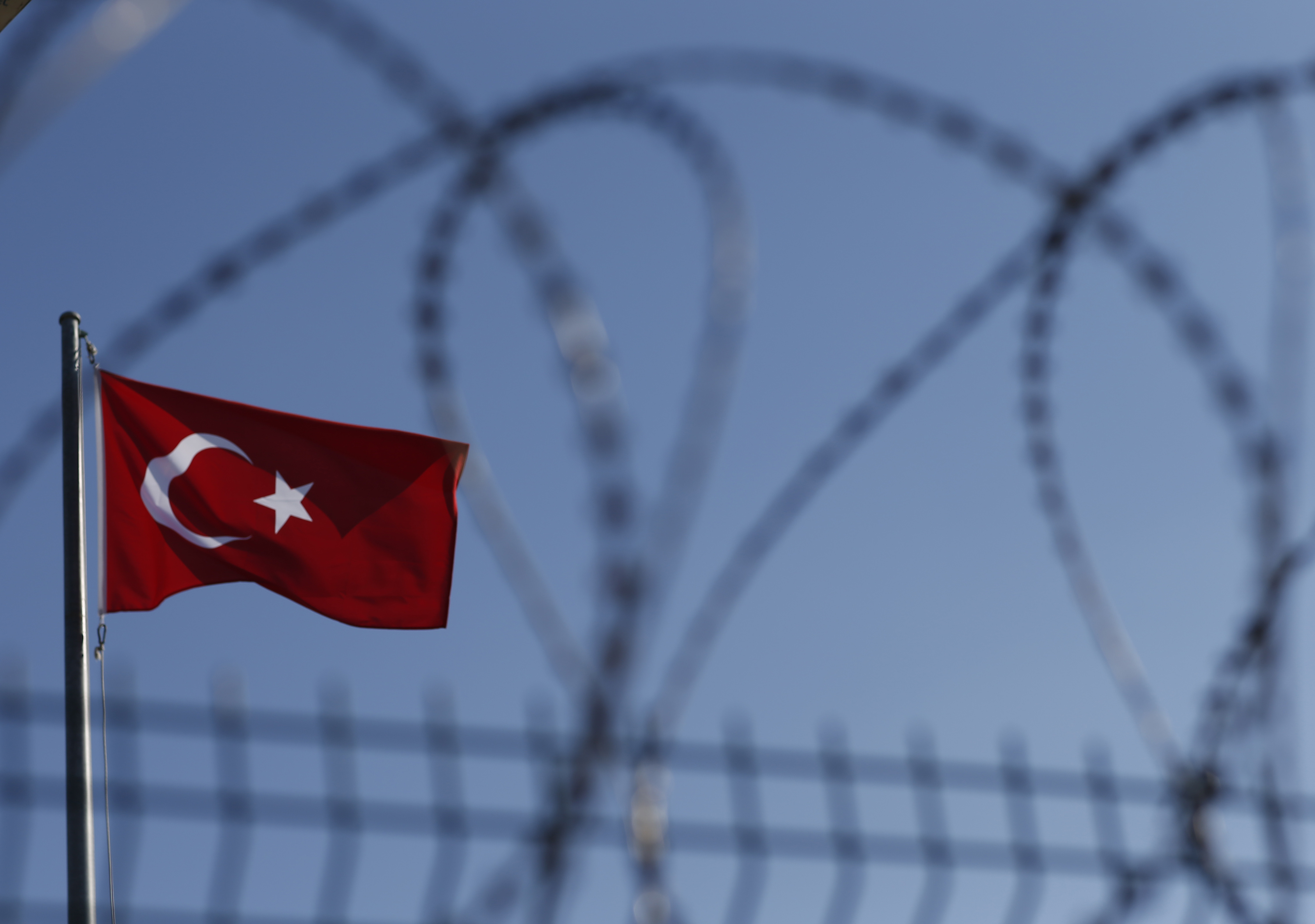 A Turkish flag fies in blue skies near a razor wire topped border fence