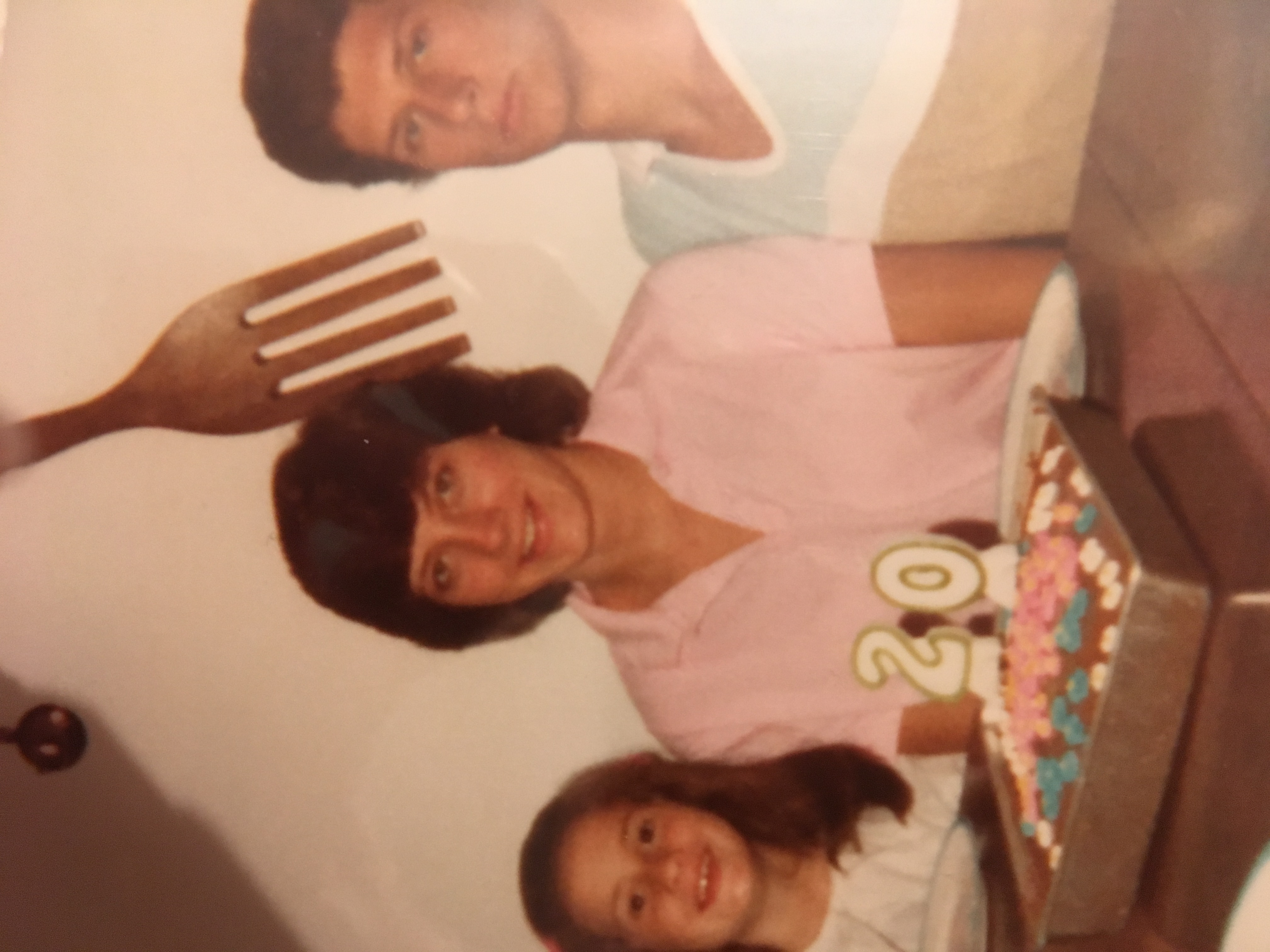 A photo of three people, a woman in the middle with a cake and a candle in the shape of the number 20 in front of her with a man sitting on the right side of the photo and a child sitting on the left.