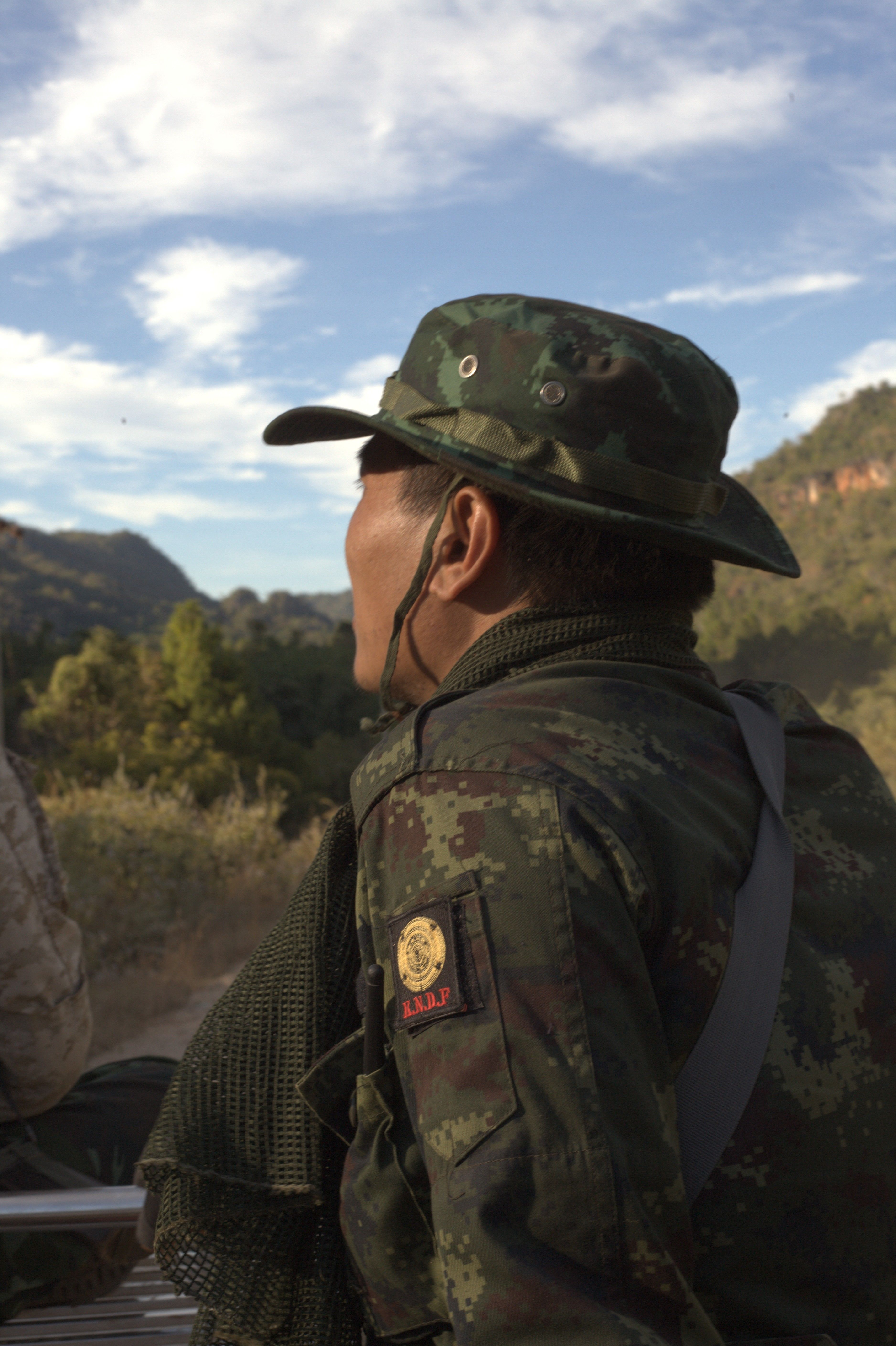 A side profile of Khun Nan Nan seen from behind wearing his green army uniform, with green mountains in the background.