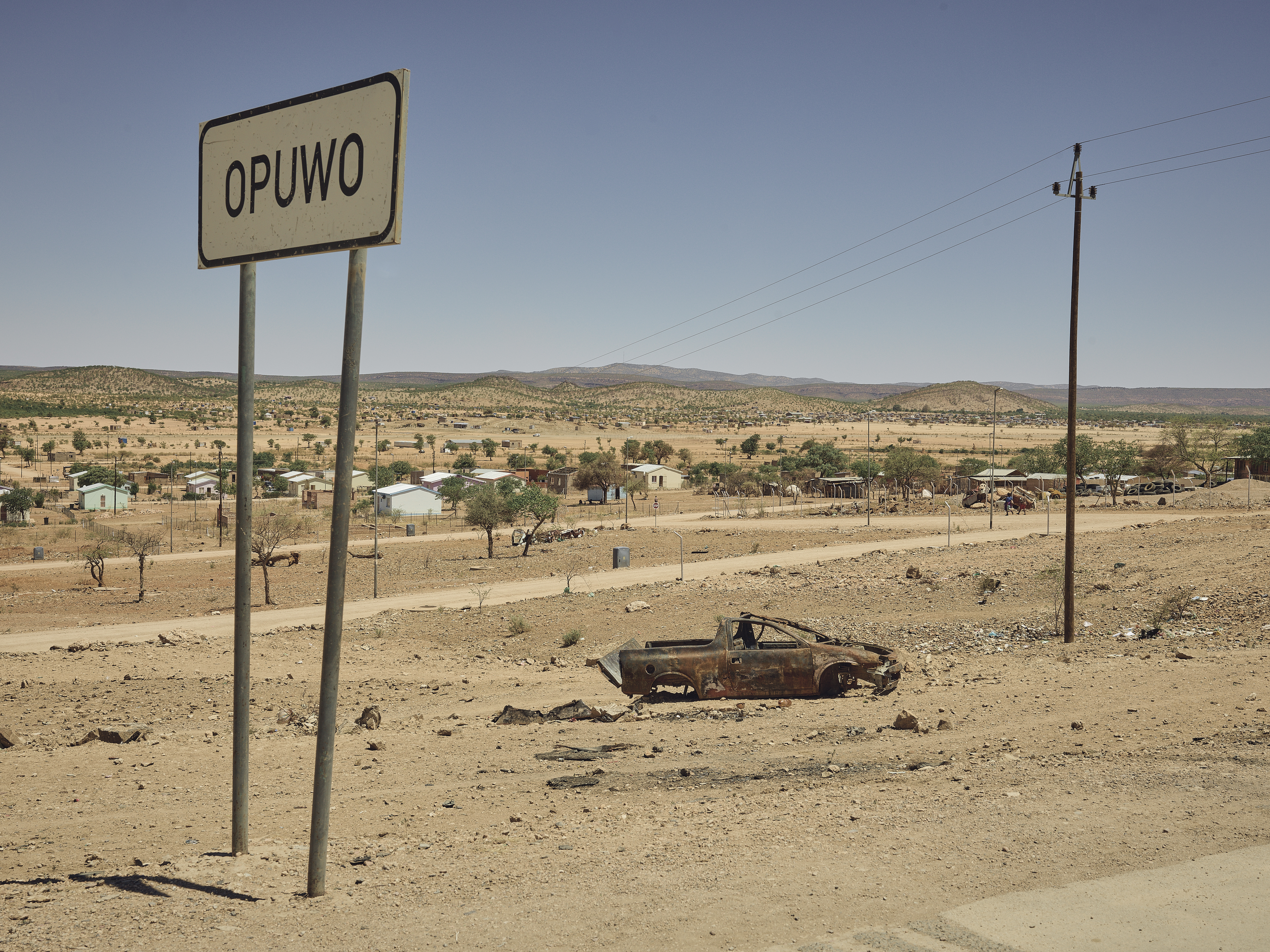 A photo of an empty road with a rusty car and a sign that says “OPUWO”.