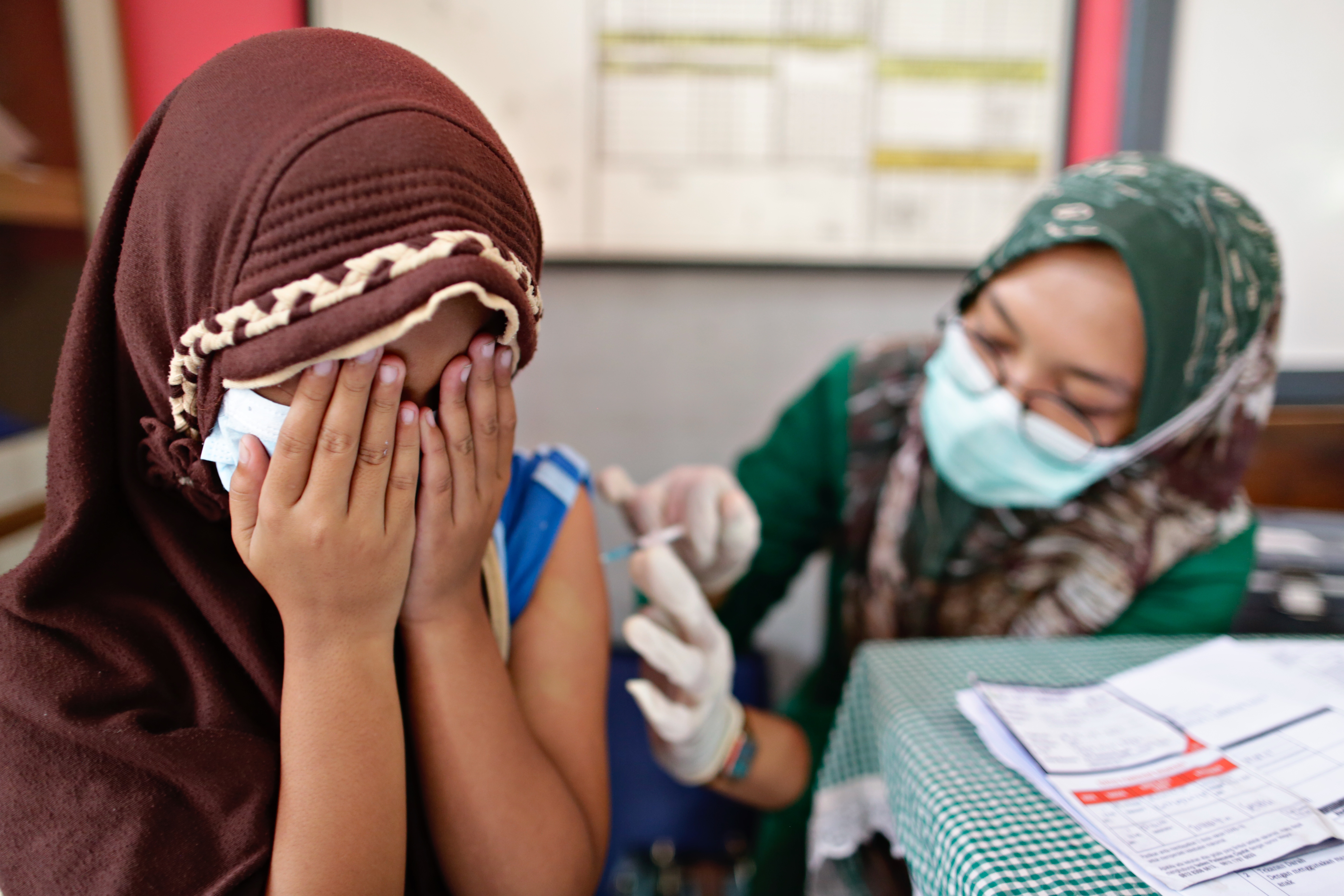 A young girl covers her face with her hands as a female nurse gives her a COVI D vaccination at school in Indonesia