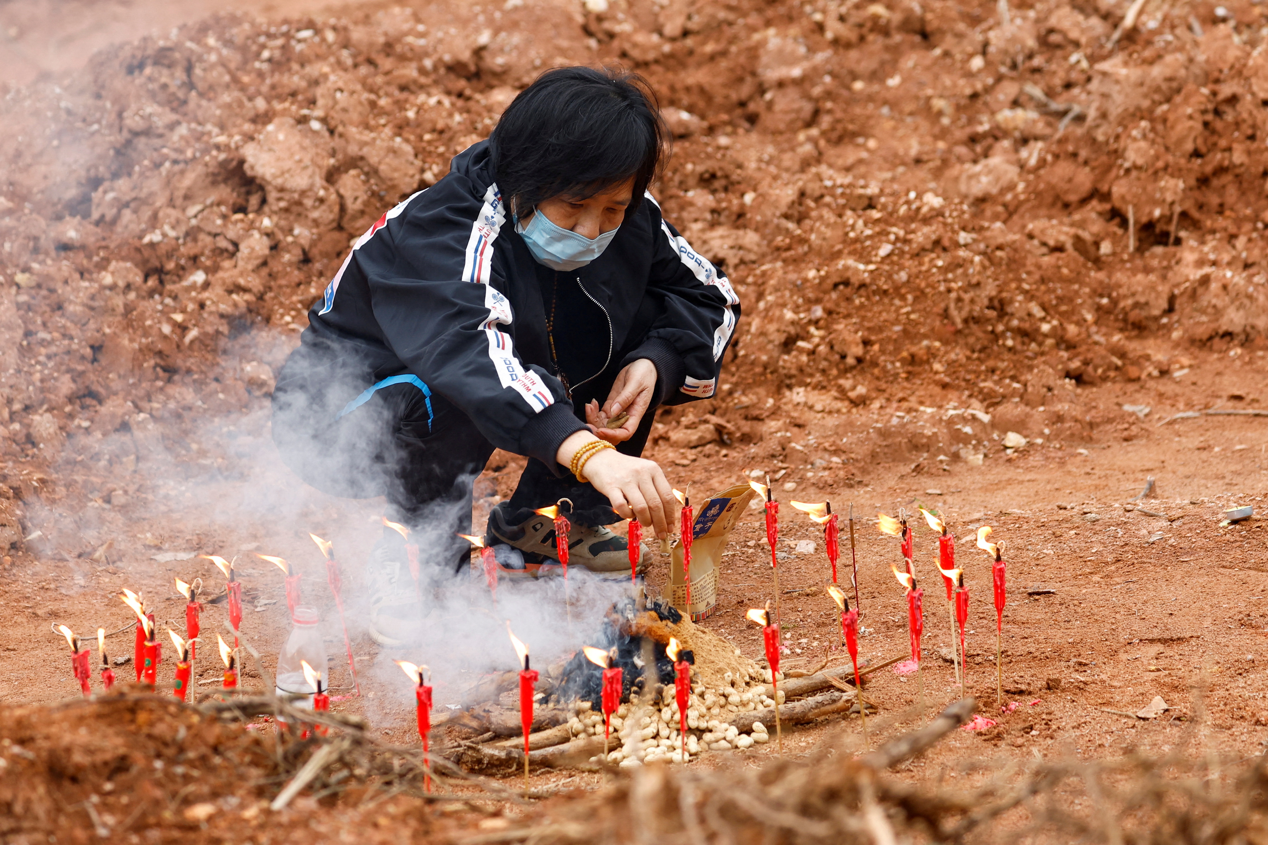 A woman squats on brown earth to light candles as part of a Buddhist ceremony for victims of the China Eastern plane crash