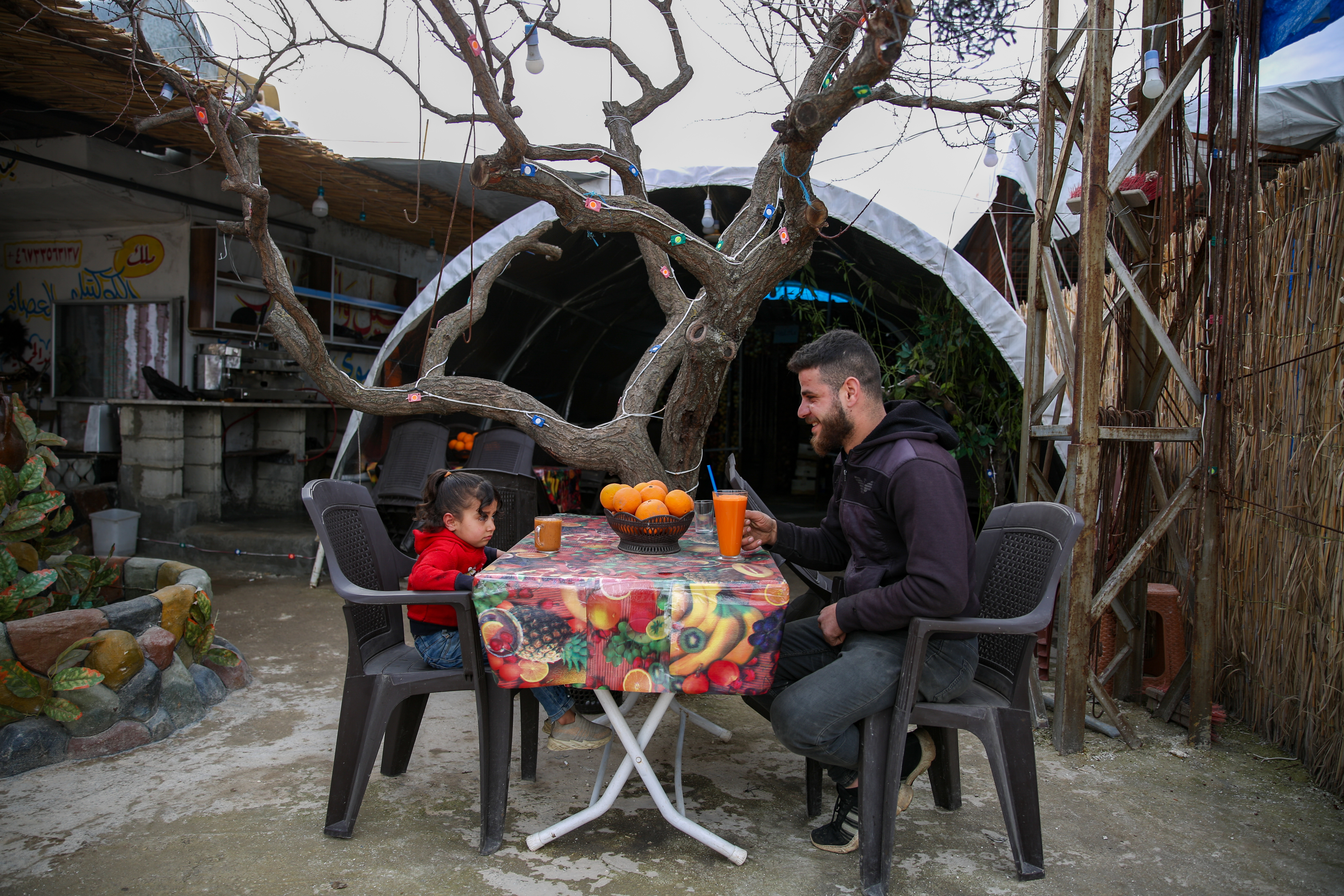 A young girl sits at a colorful table drinking juice with her older male relative. They are in a grey courtyard.