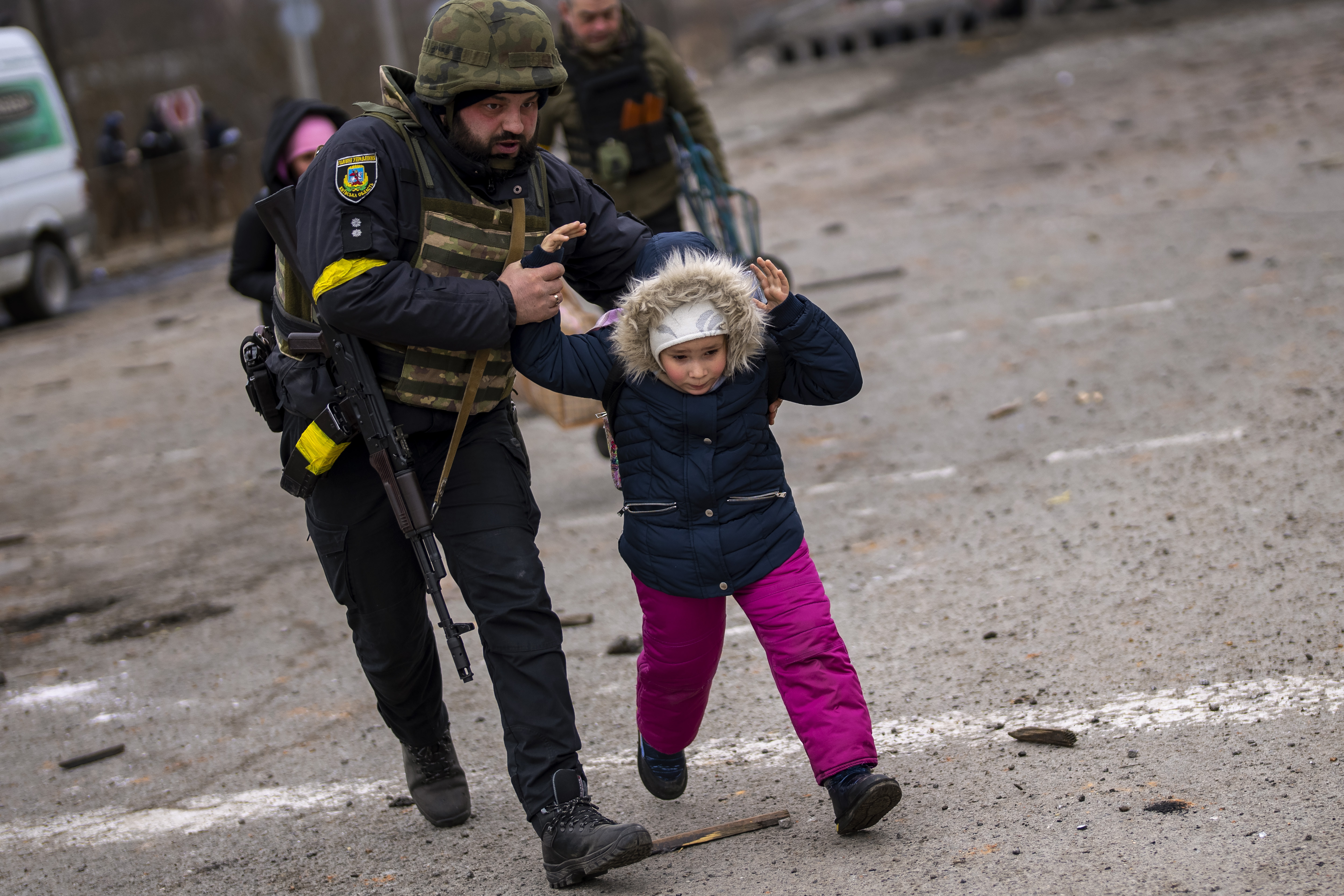 A Ukrainian police officer runs while holding a child as the artillery echoes nearby, while fleeing Irpin on the outskirts of Kyiv