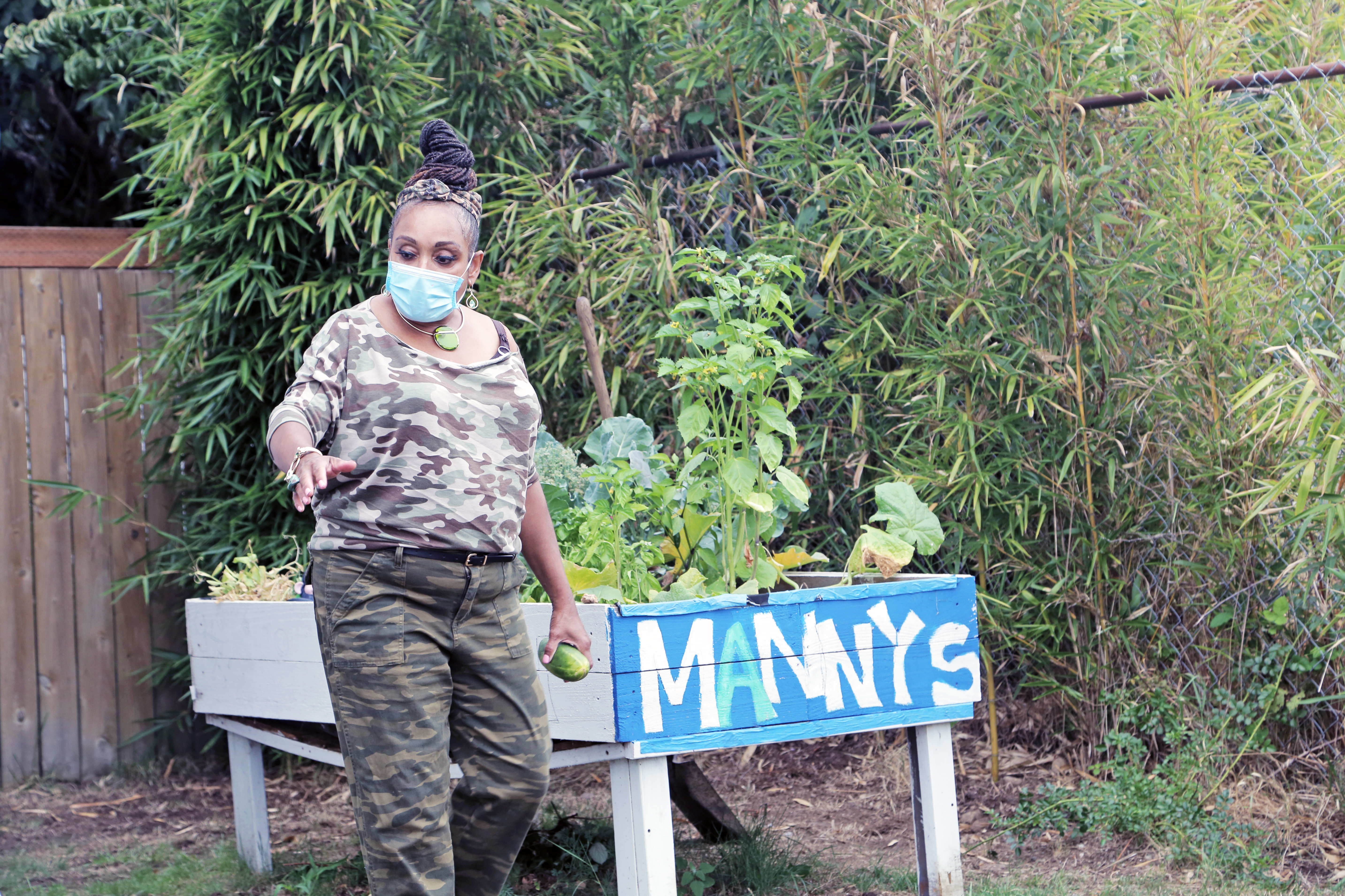 Marcia Carter, wearing trousers and a t-shirt in a camouflage print and a face mask, stands in a garden beside a blue wooden sign with white painted letters spelling Manny's