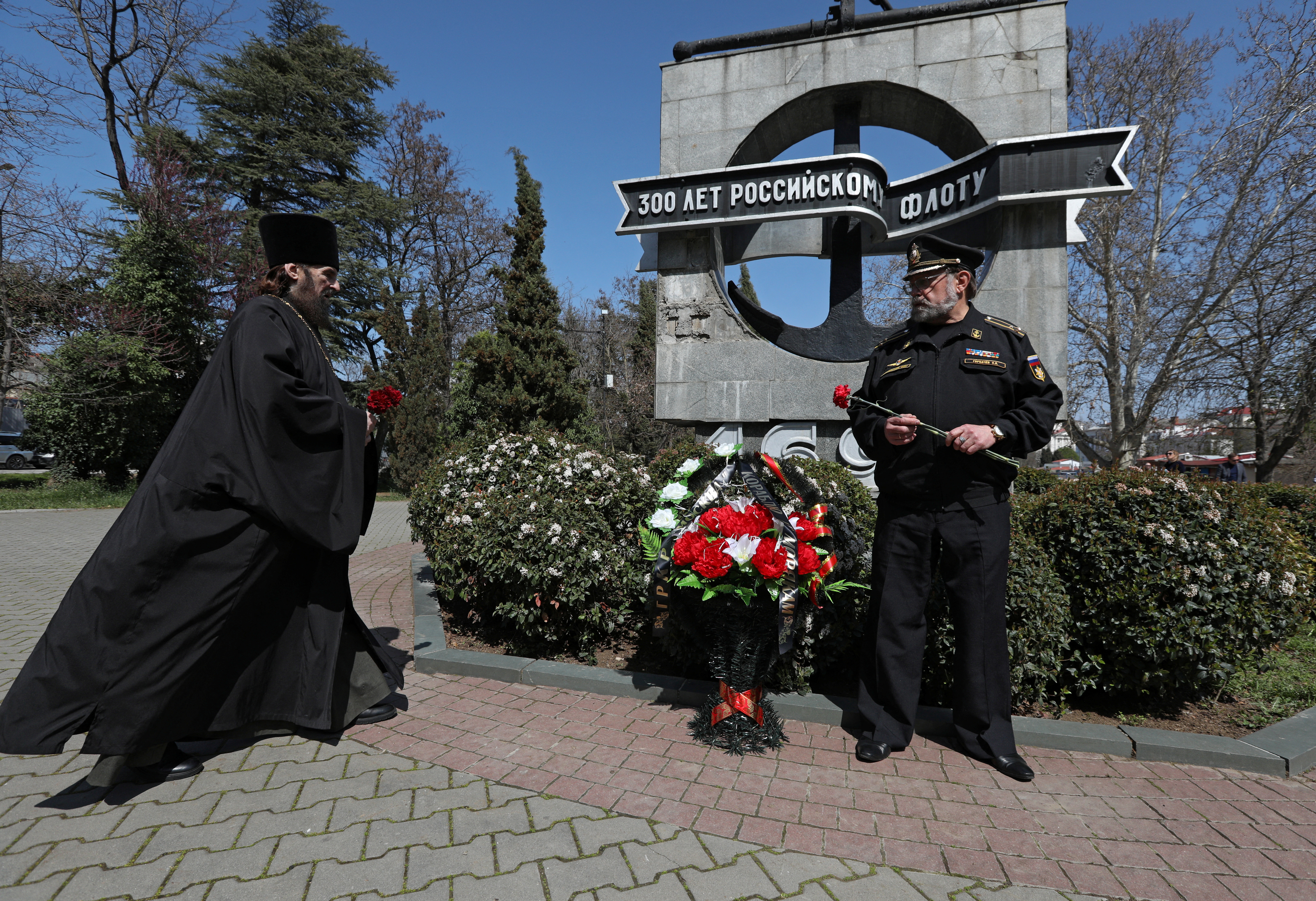 A Russian orthodox priest in black robes with a Black Sea Fleet veteran at a ceremony in honour of the Moskva in front of a Soviet-style sculpture strewn with flowers