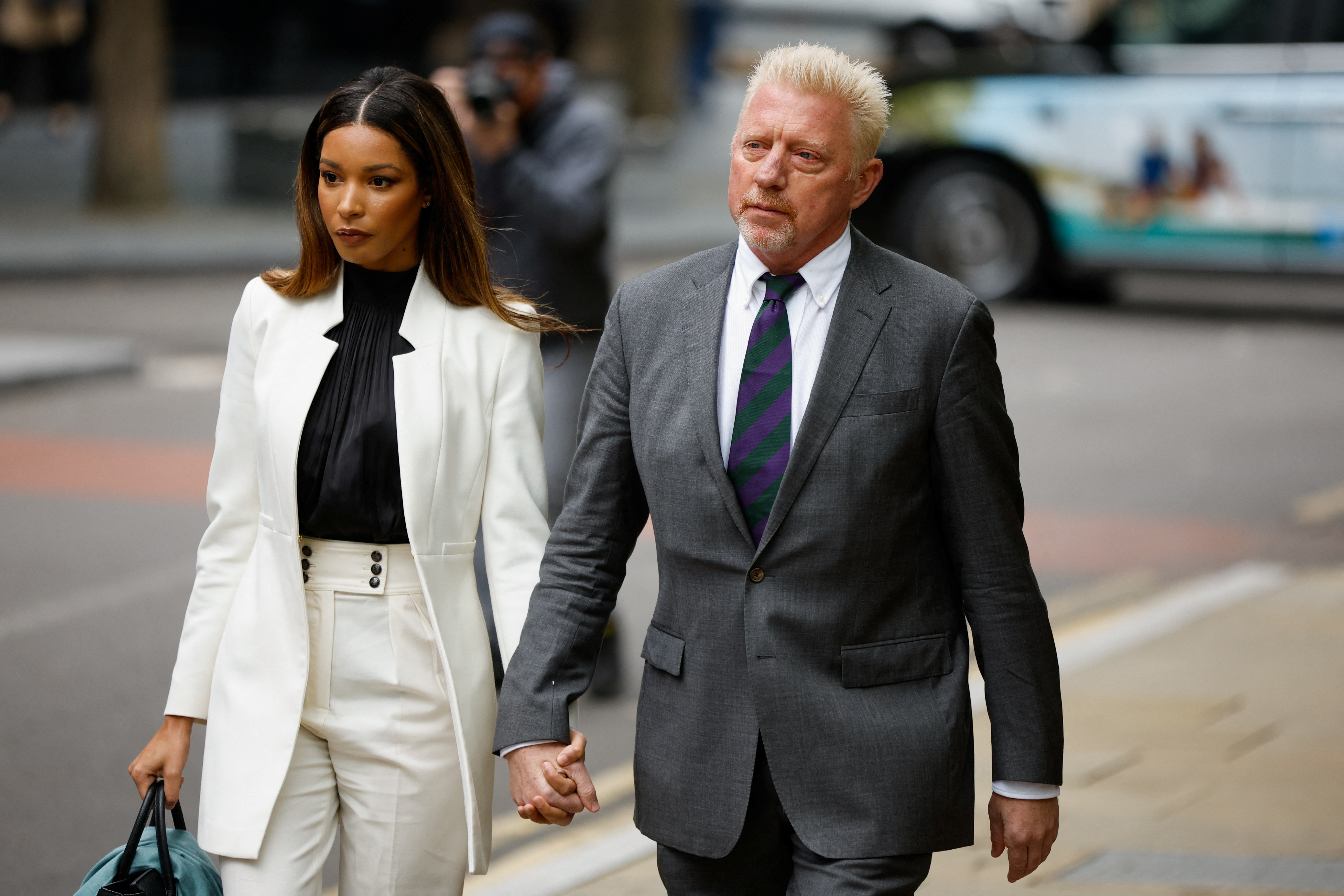 Boris Becker in a grey suit and tie walks to court with his partner partner Lilian de Carvalho Monteiro in white trouser suit and black shirt