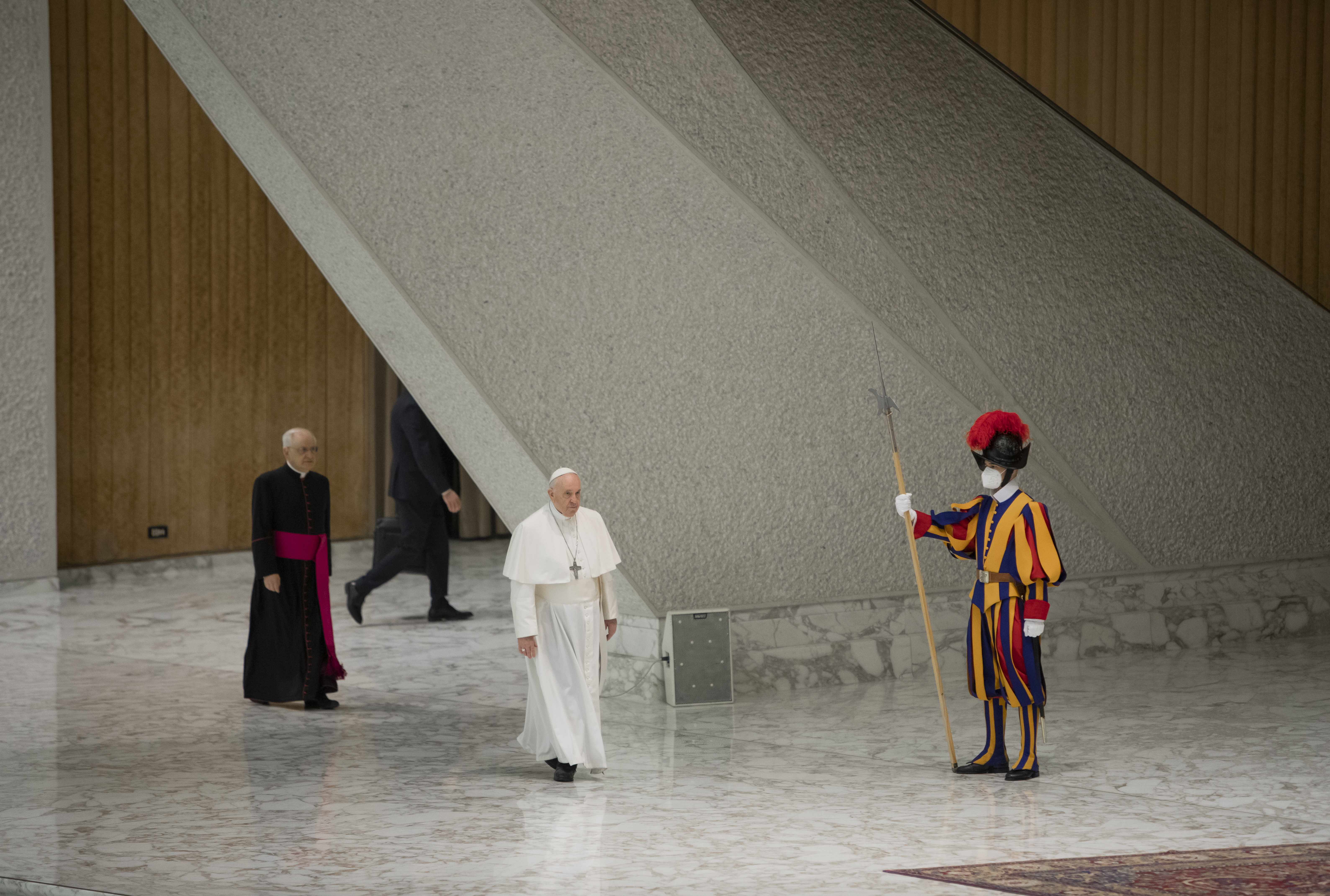 A photo of Pope Francis walking with a bishop behind him and a security guard standing in front of him.