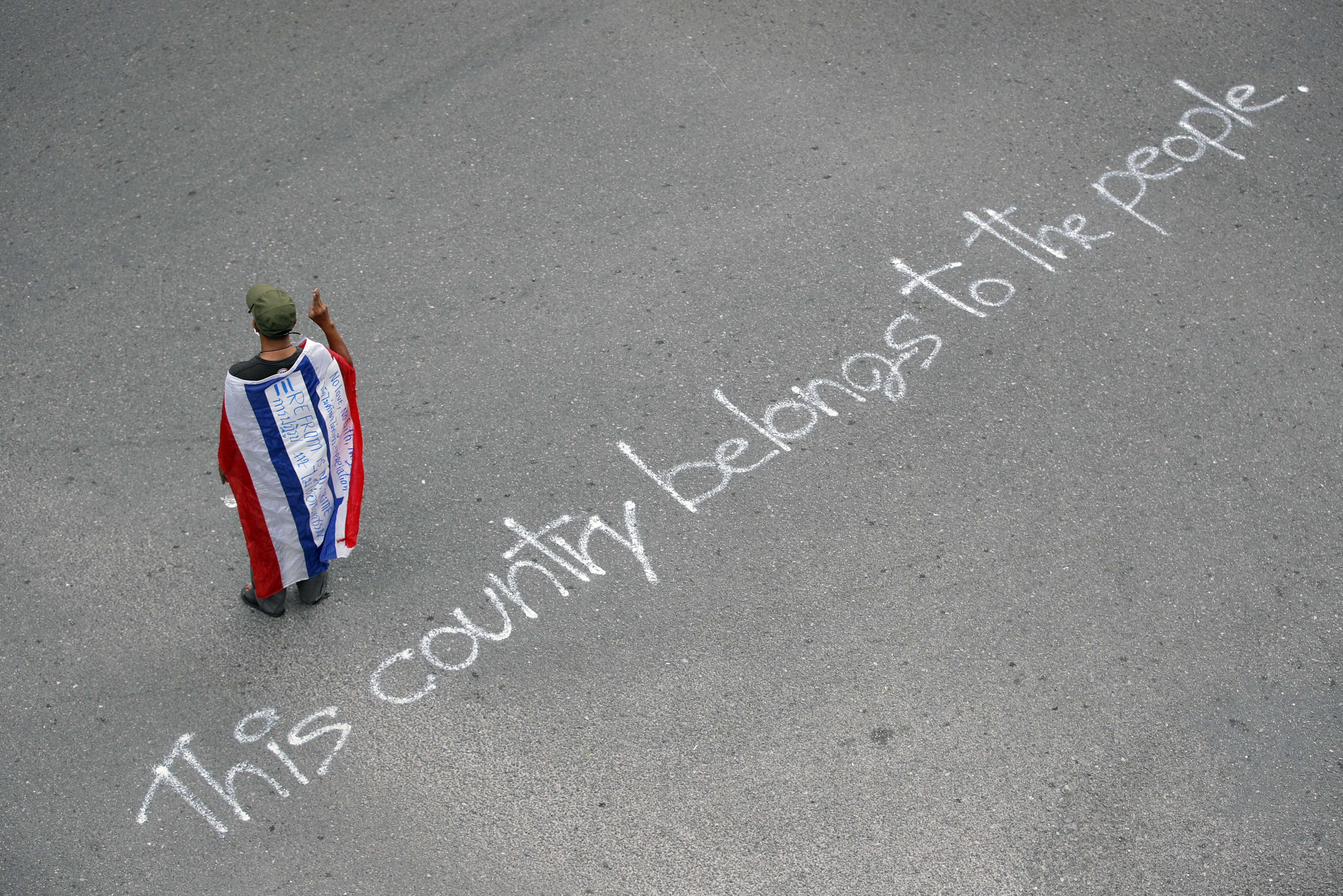 A protester draped in a Thai flag stands on a road in Bangkok where demonstrators have written 'this country belongs to the people" in white across the grey tarmac.