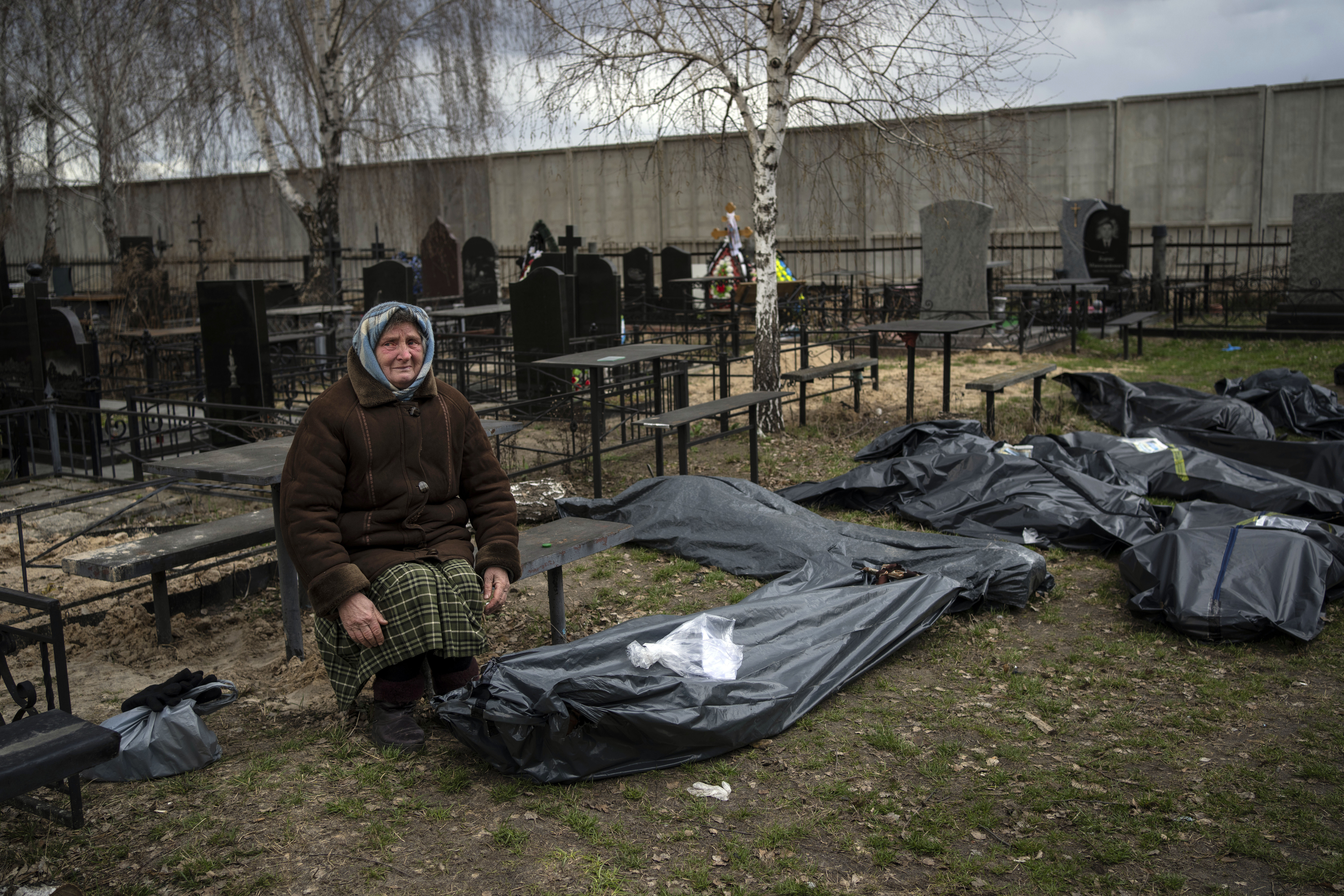 Elderly woman kneels next to corpses wrapped in body bags