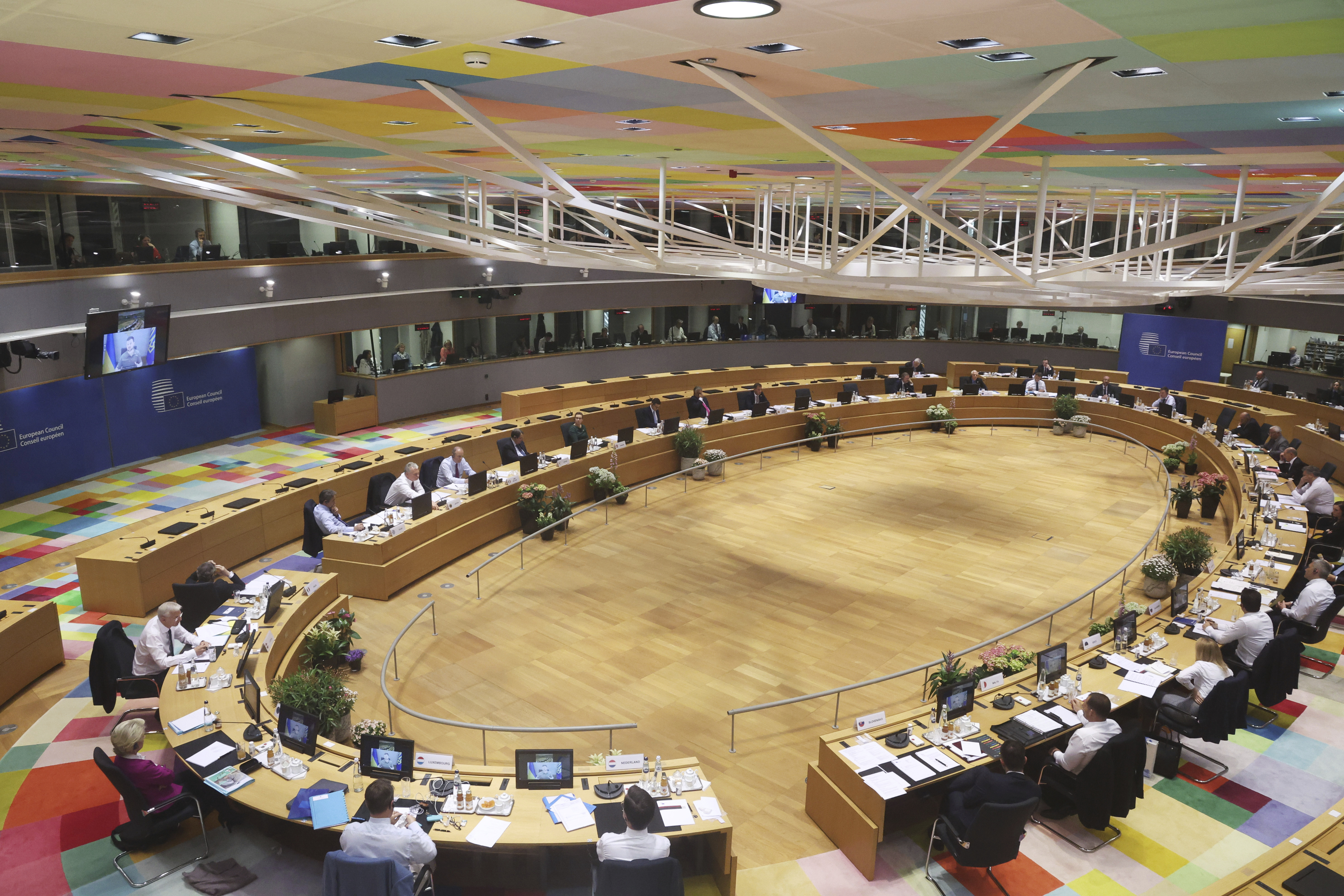 A wide view of the meeting hall and delegates sitting around the oval shaped table as Volodymyr Zelenskyy speaks via video link and is seen on a large screen to one side of the hall