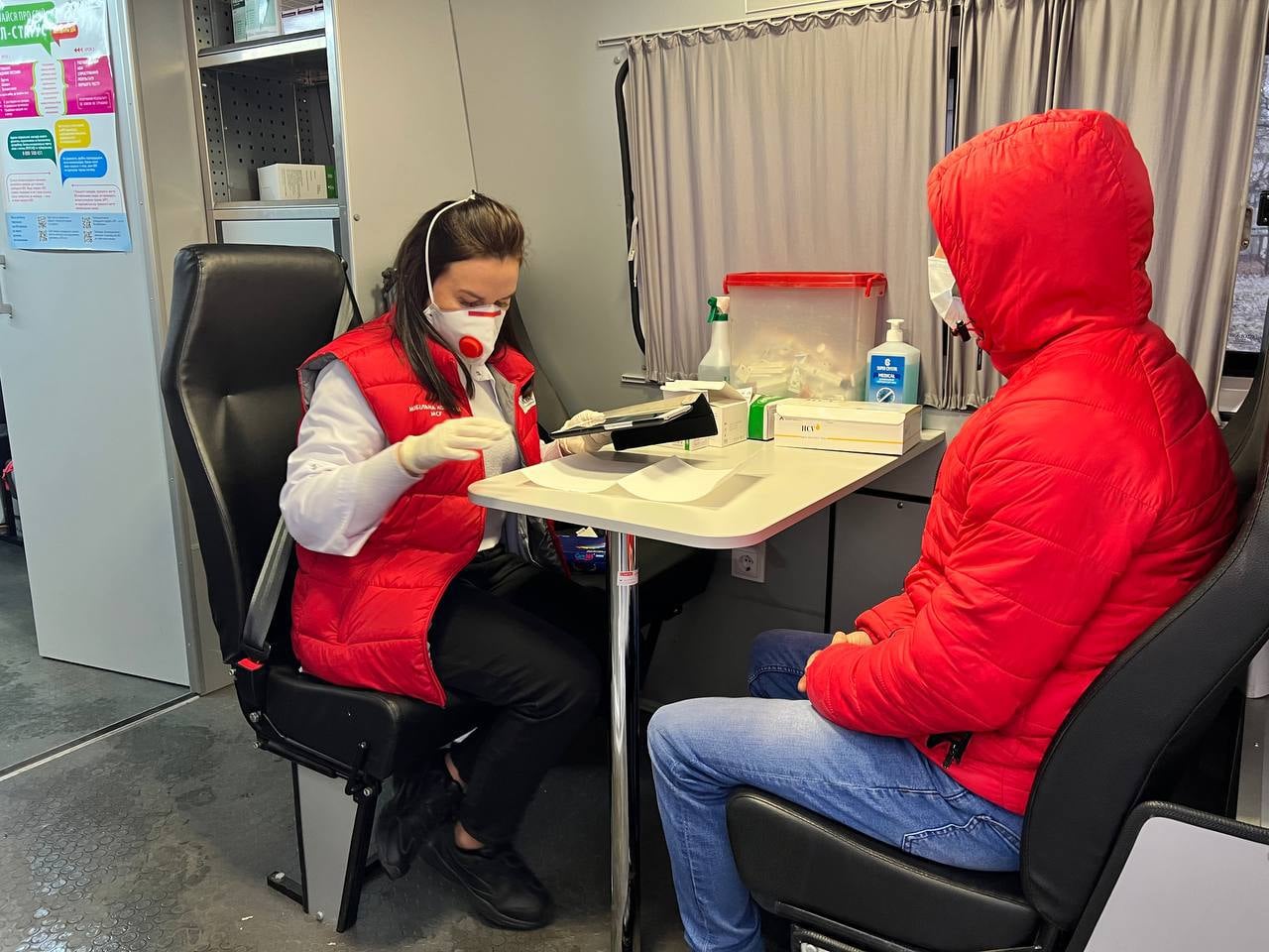 A photo of two people sitting on opposite ends of a small table, the woman is looking at a piece of paper at a desk.