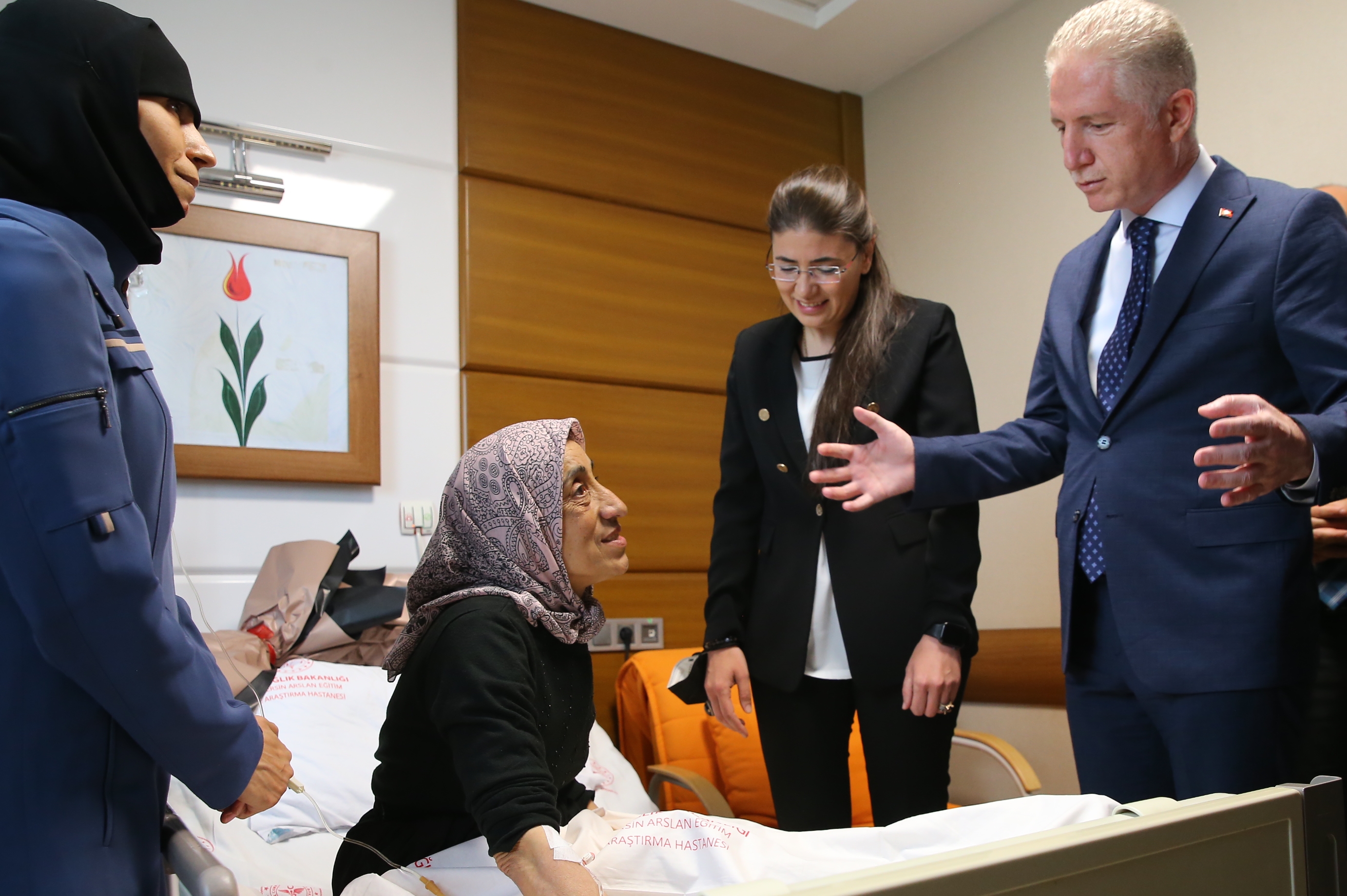 A man in a suit greets an elderly woman on a hospital bed as two people look on