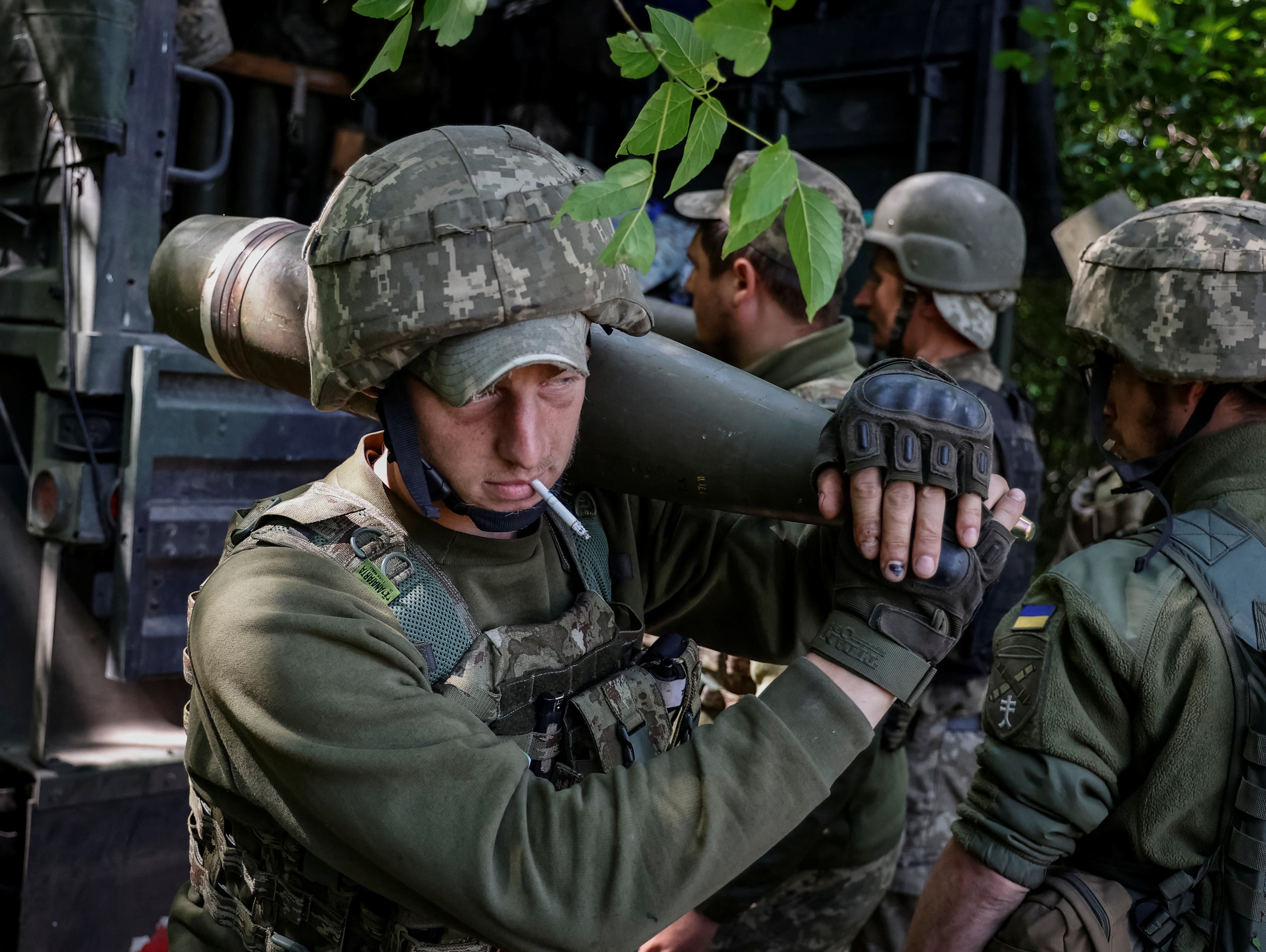 A Ukrainian service member holding a shell for an M777 Howitzer on his shoulder with other Ukrainian service members preparing shells for a M777 Howitzer in the background.