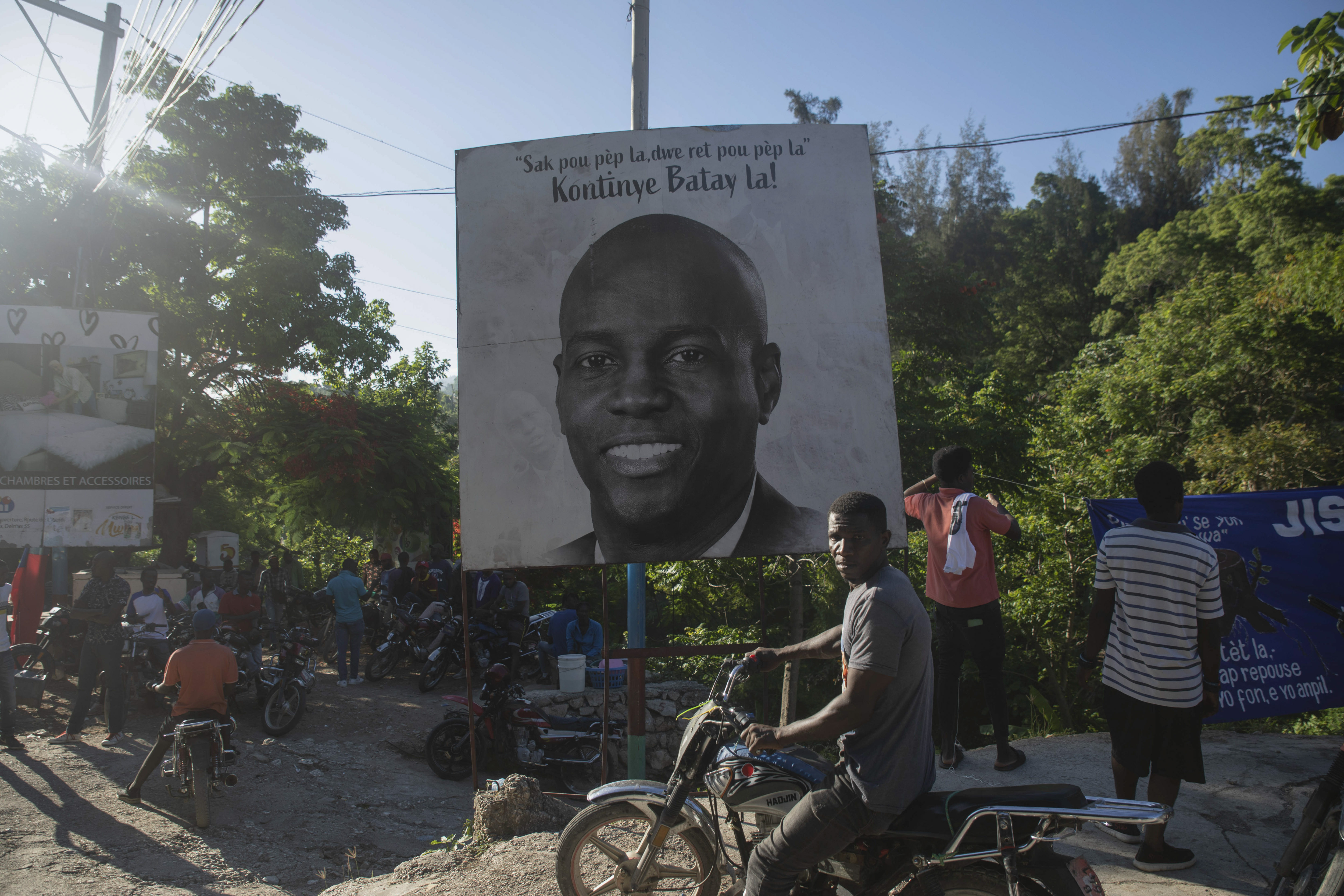 Man on a motorbike in front of the poster looks at the camera. There is a crowd of men on motorbikes next to the poster