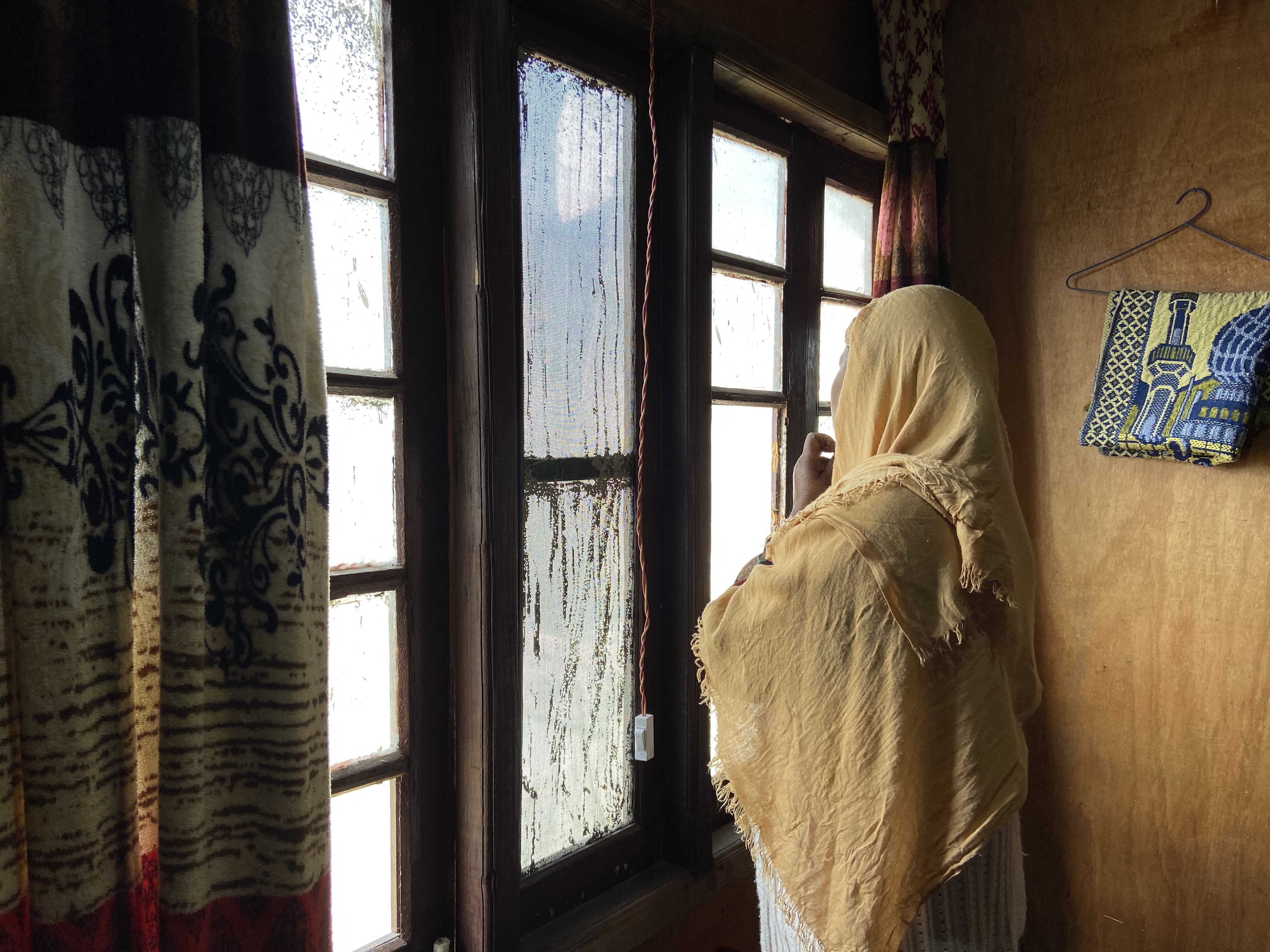A photo of a woman looking outside through a window indoors.