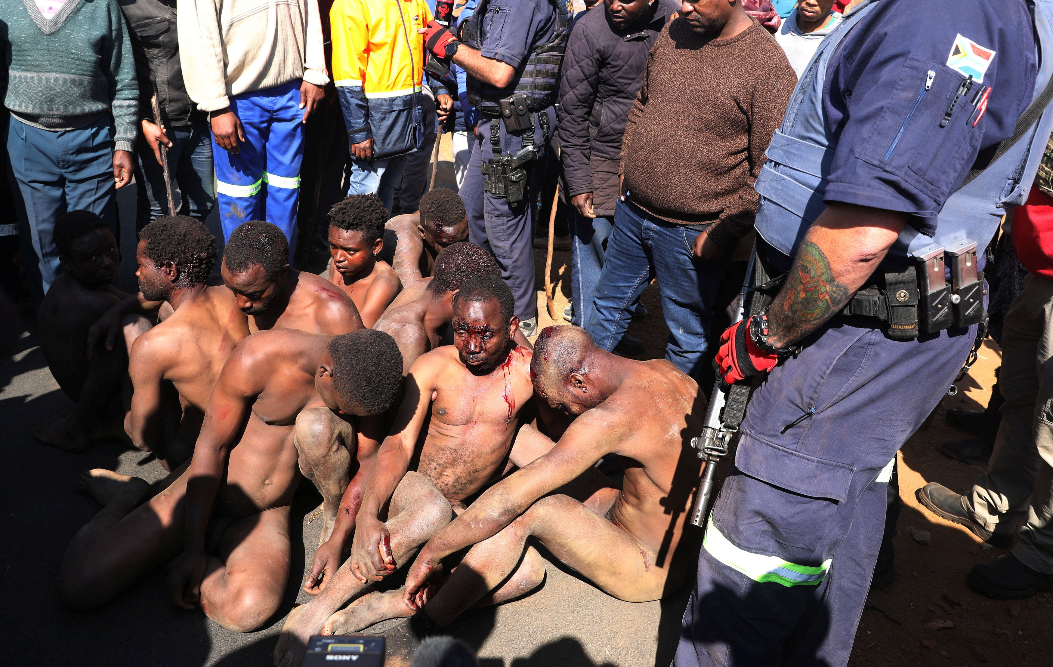 Police officers and locals stand next to alleged illegal miners known as zama-zamas after residents shut down their camps over the recent Krugersdorp mass rape.