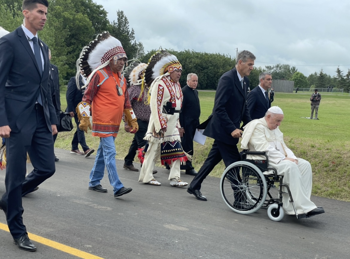 A photo of someone pushing the pope in a wheelchair with people all around.