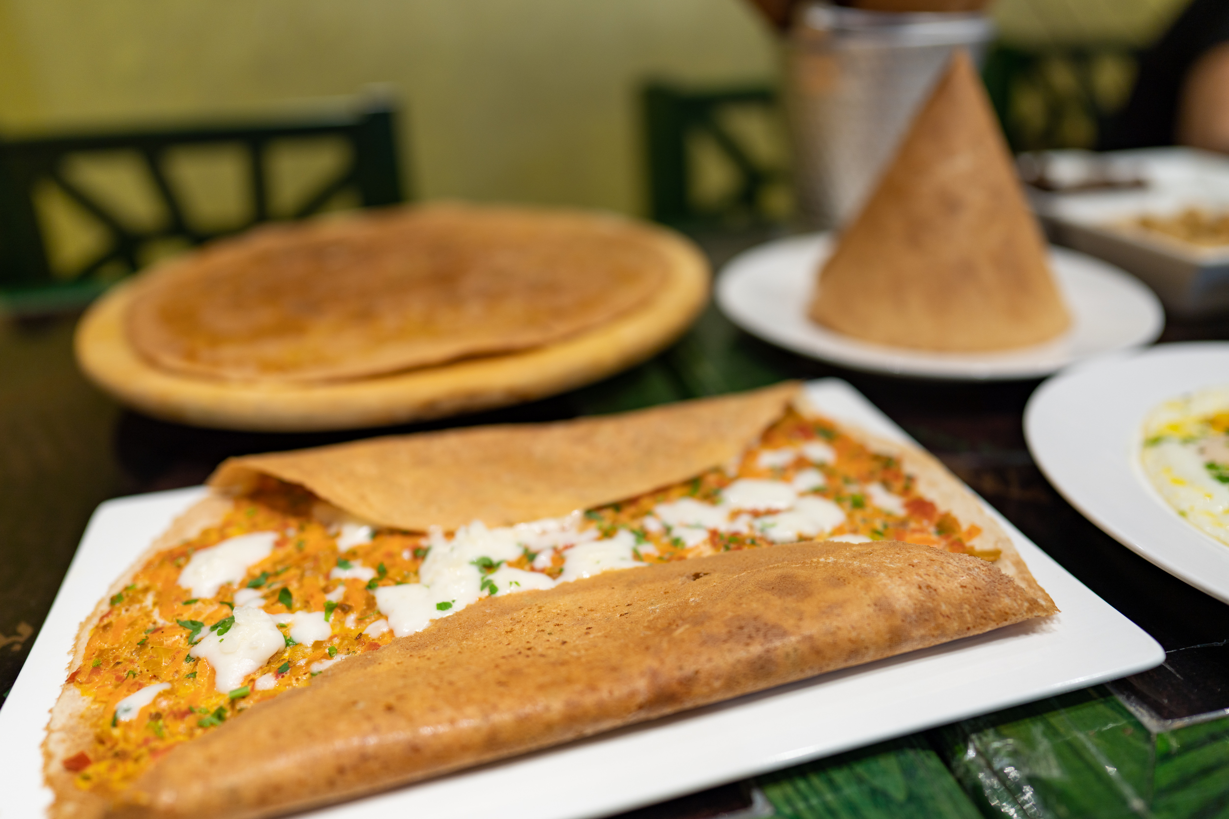 In the foreground is a regag bread with its sides folded in part-way to show its egg, veggie and cheese filling. Background is a regag cone and a flat shumeelah regag