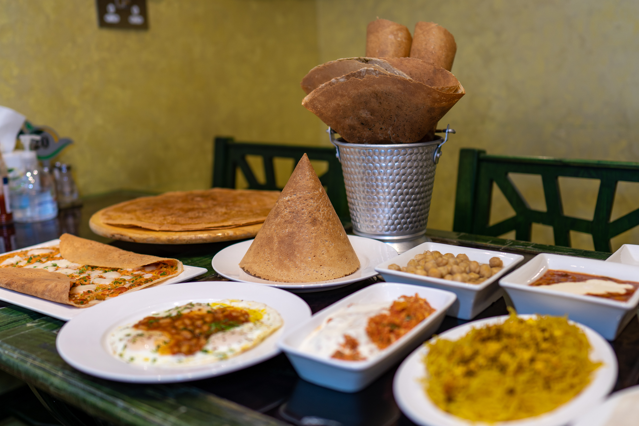 Another view of the green table with breakfast food on it. Prominent in it is a plate with three fried eggs topped with spicy baked beans and a sprinkle of copped herbs
