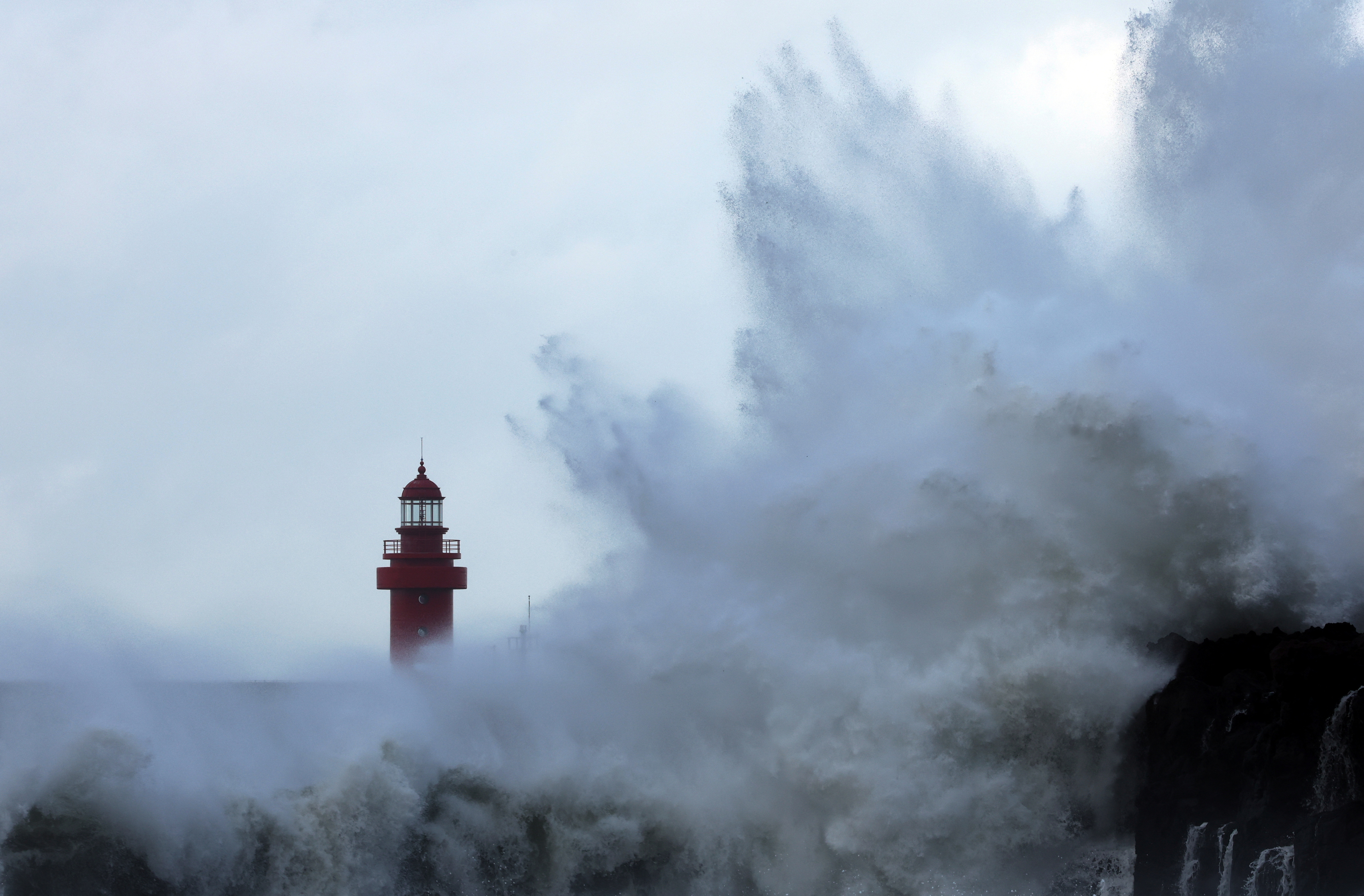 A giant wave crashes against the coast of Jeju island with the top of a lighthouse visible behind