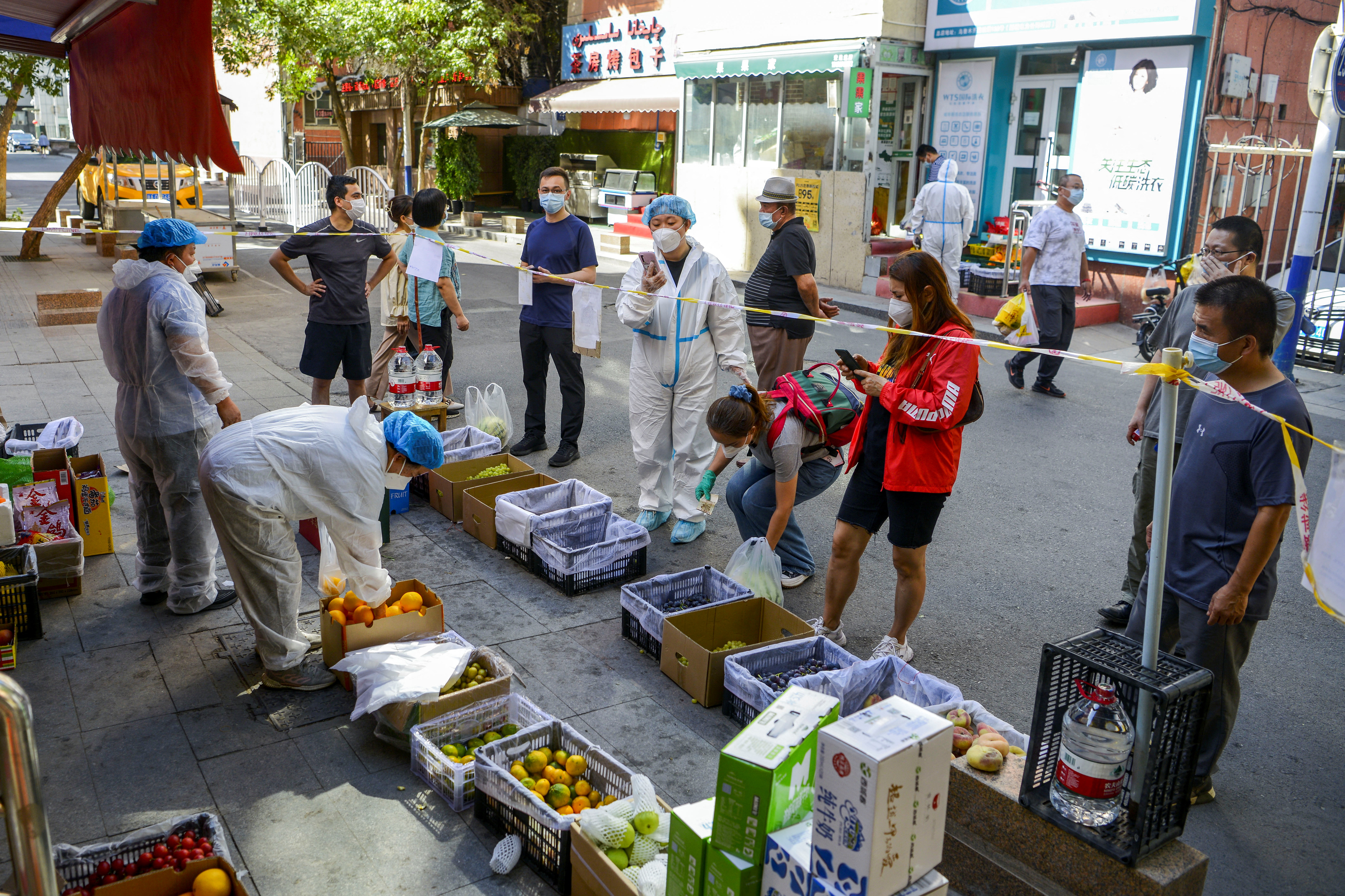 People queue behind a tape in masks at a fruit stall in Urumqi with the two stallholders in hazmat suits.