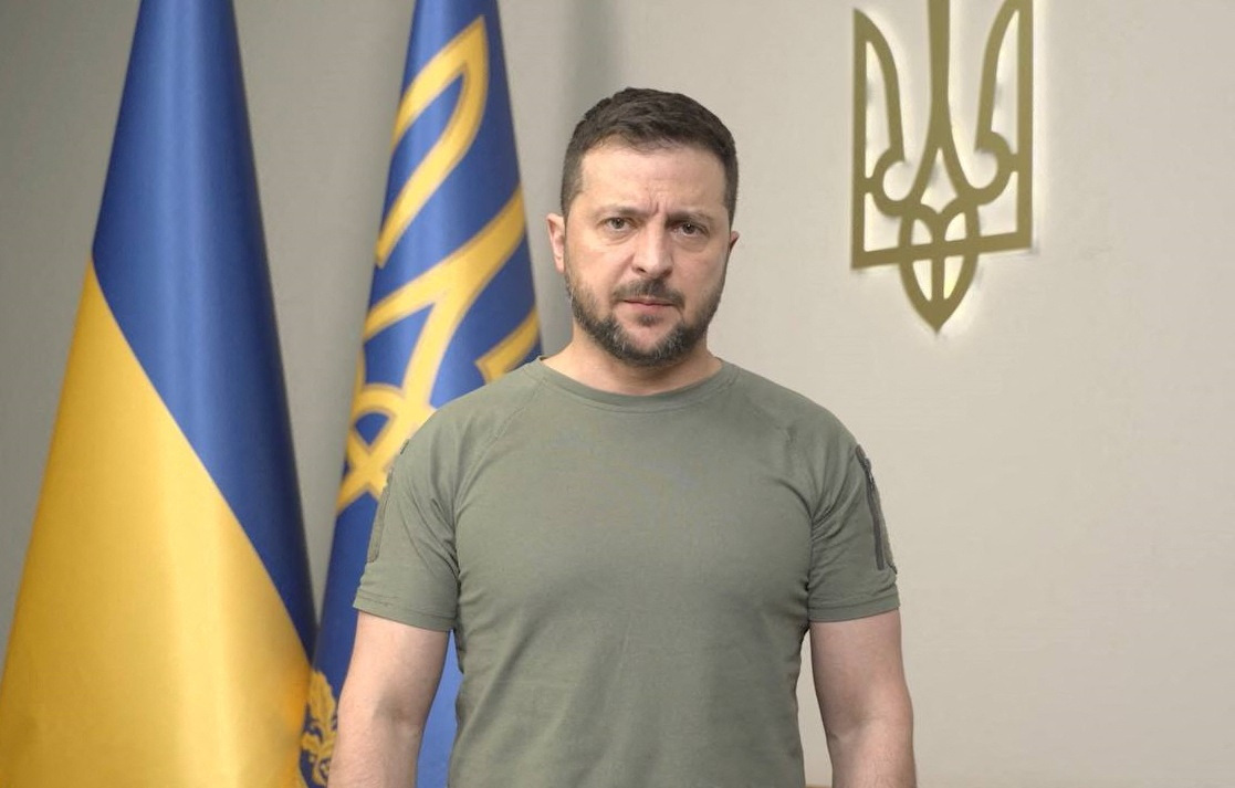 Zelenskyy in dark green shirt with Ukrainian flags behind him