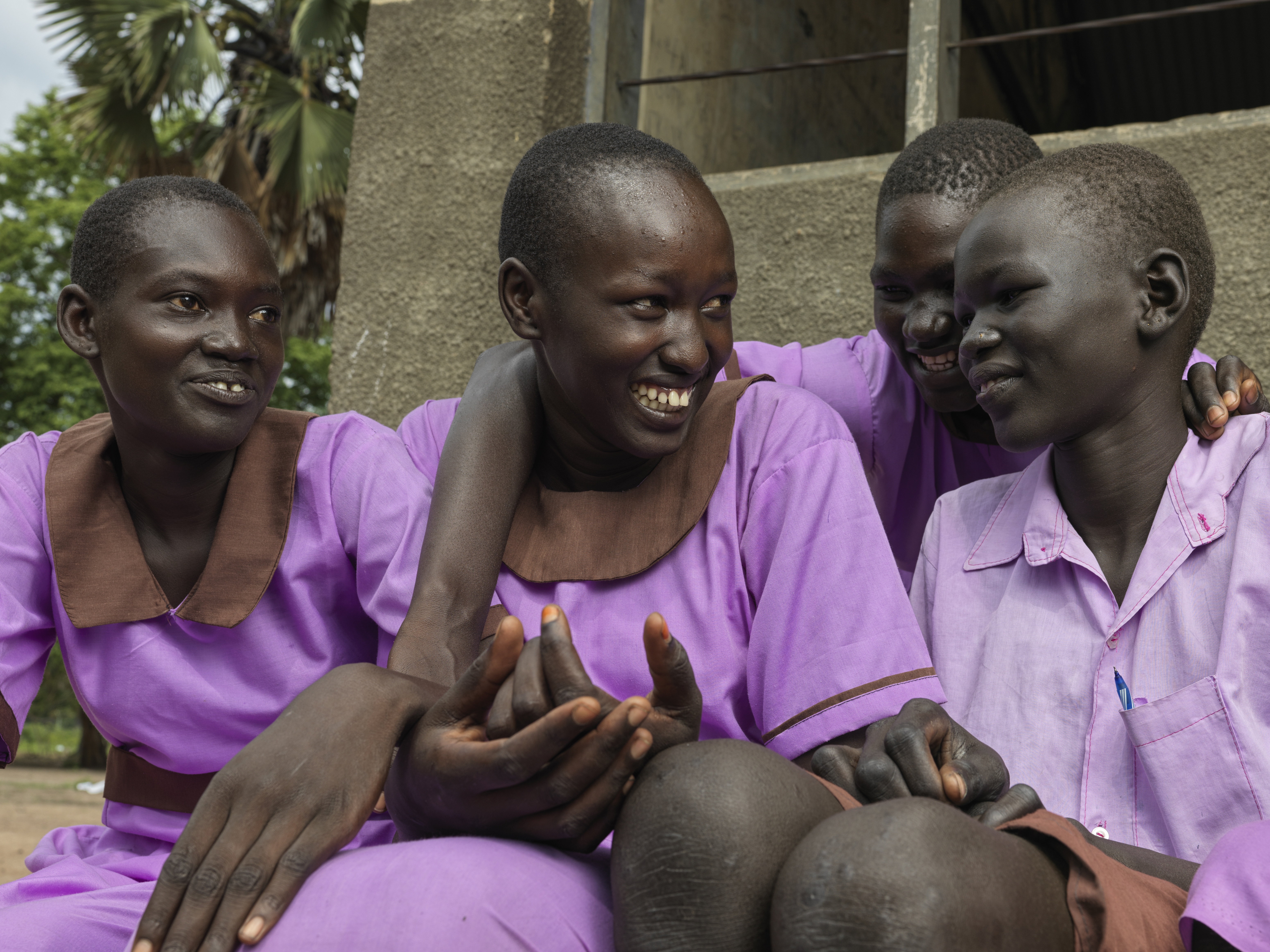 A photo of four students sitting and talking and laughing together, three are next to each other and one is in the back with their arms around two of the ones in the front..