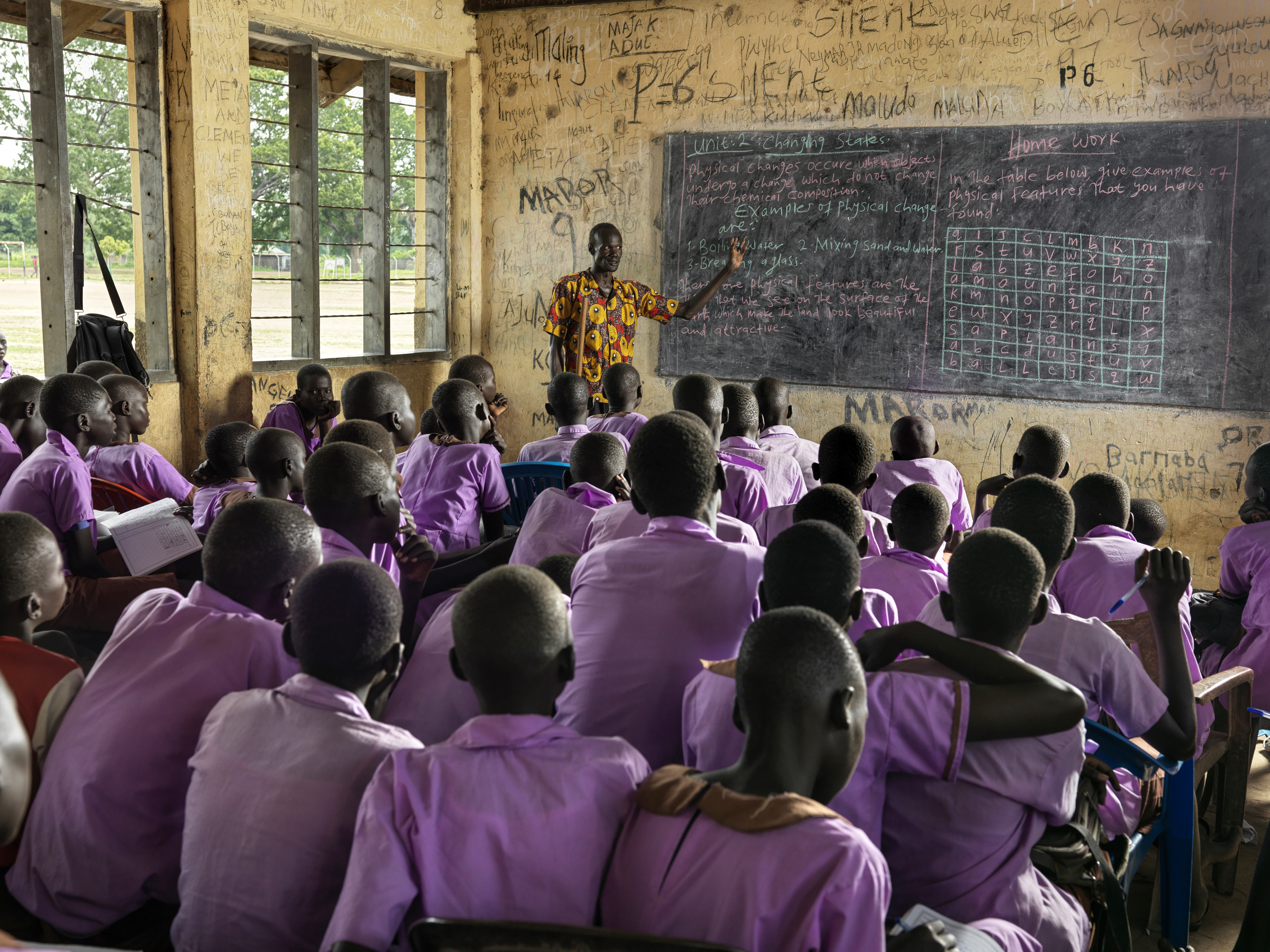 A photo of a classroom of students with a teacher in the front pointing to something on a blackboard.