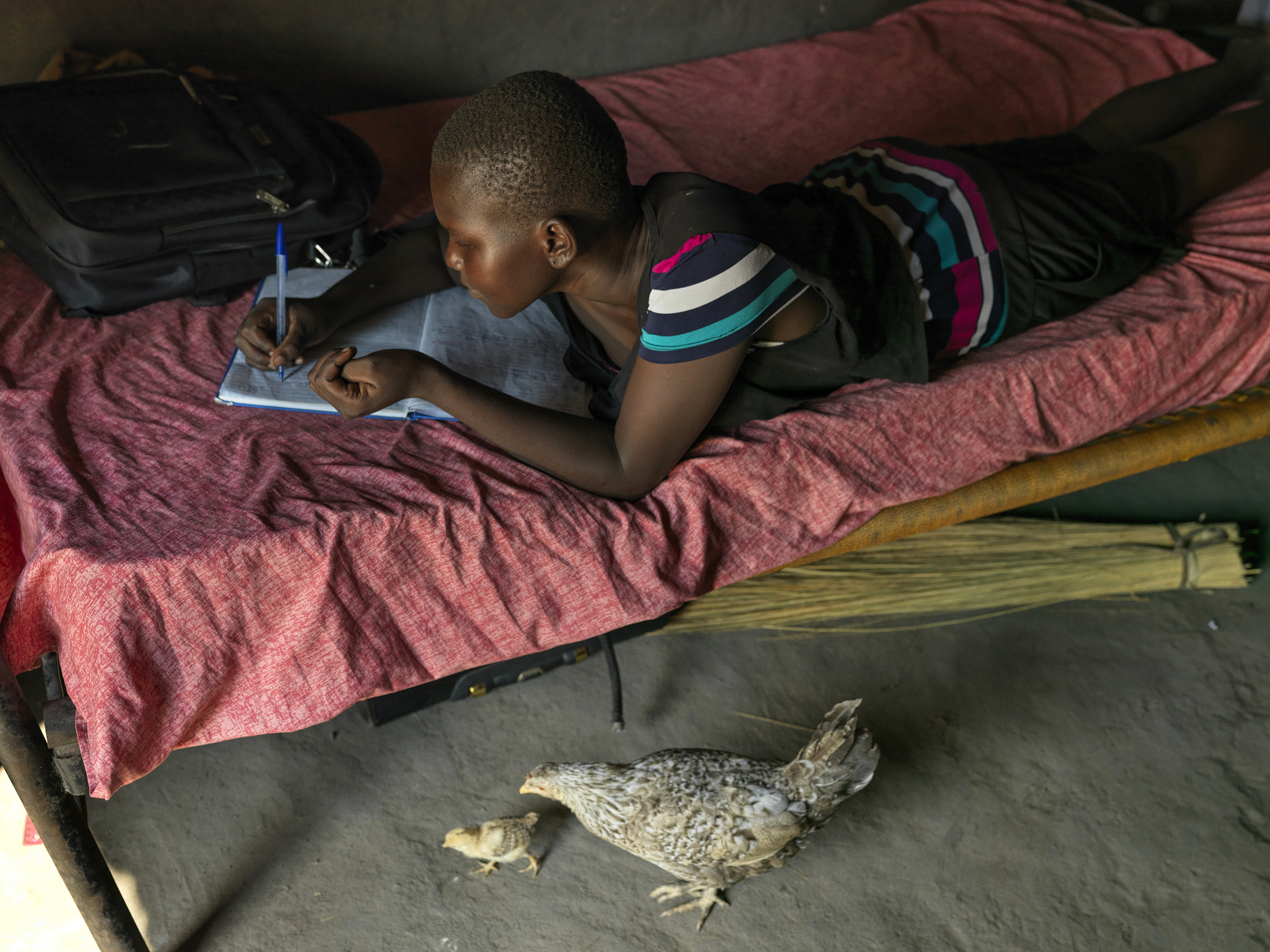A photo of Grace laying on her stomach writing in a notebook on her bed with a chicken and a little chick under her bed.