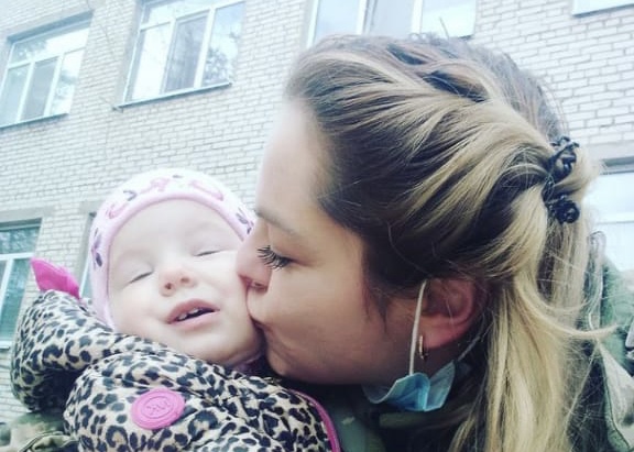 Anna holding her daughter Milena, who is wearing a pink hat, and kissing her cheek.