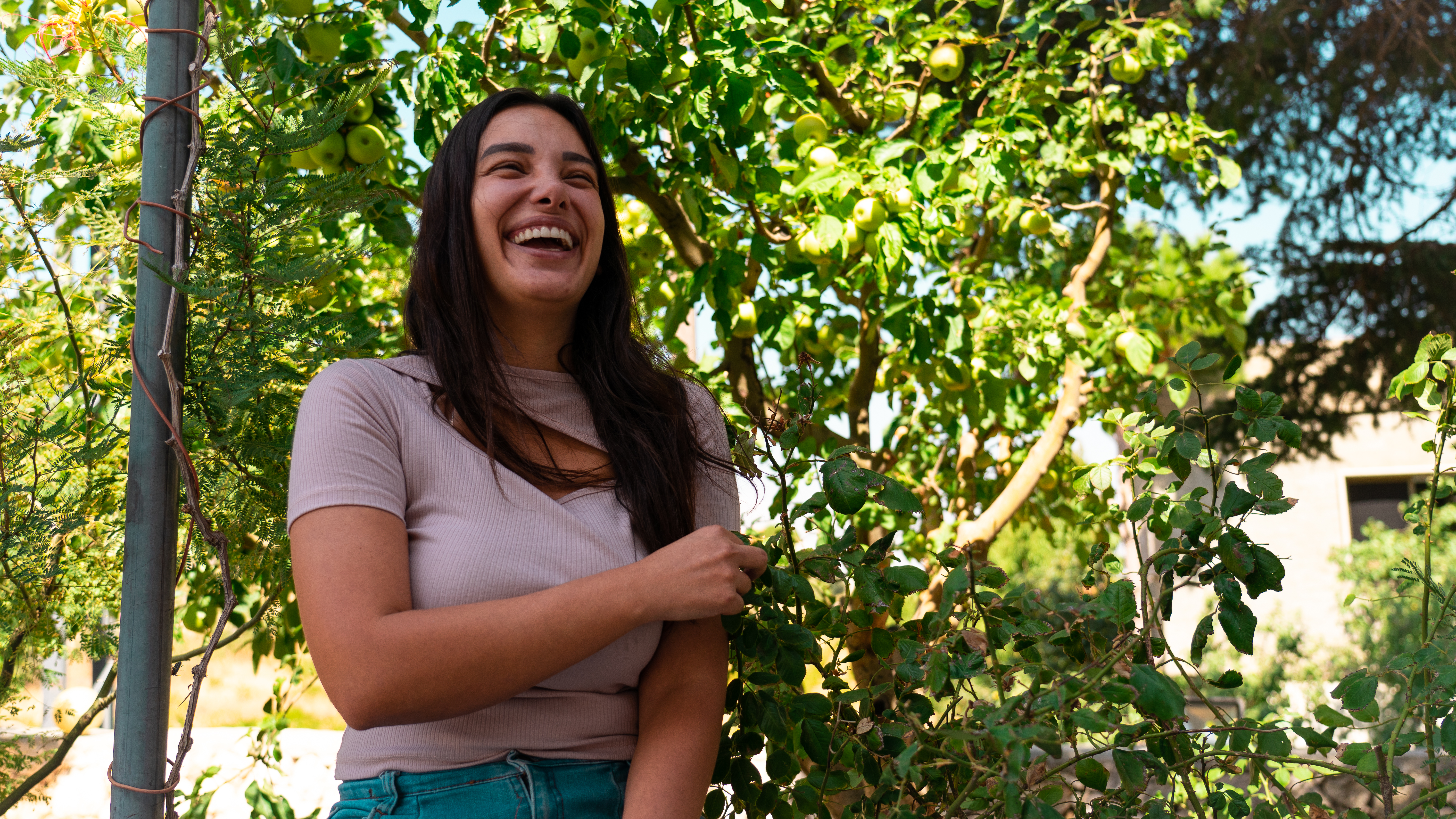 Sali Hafiz stands laughing and holding her left arm in front of an apple tree