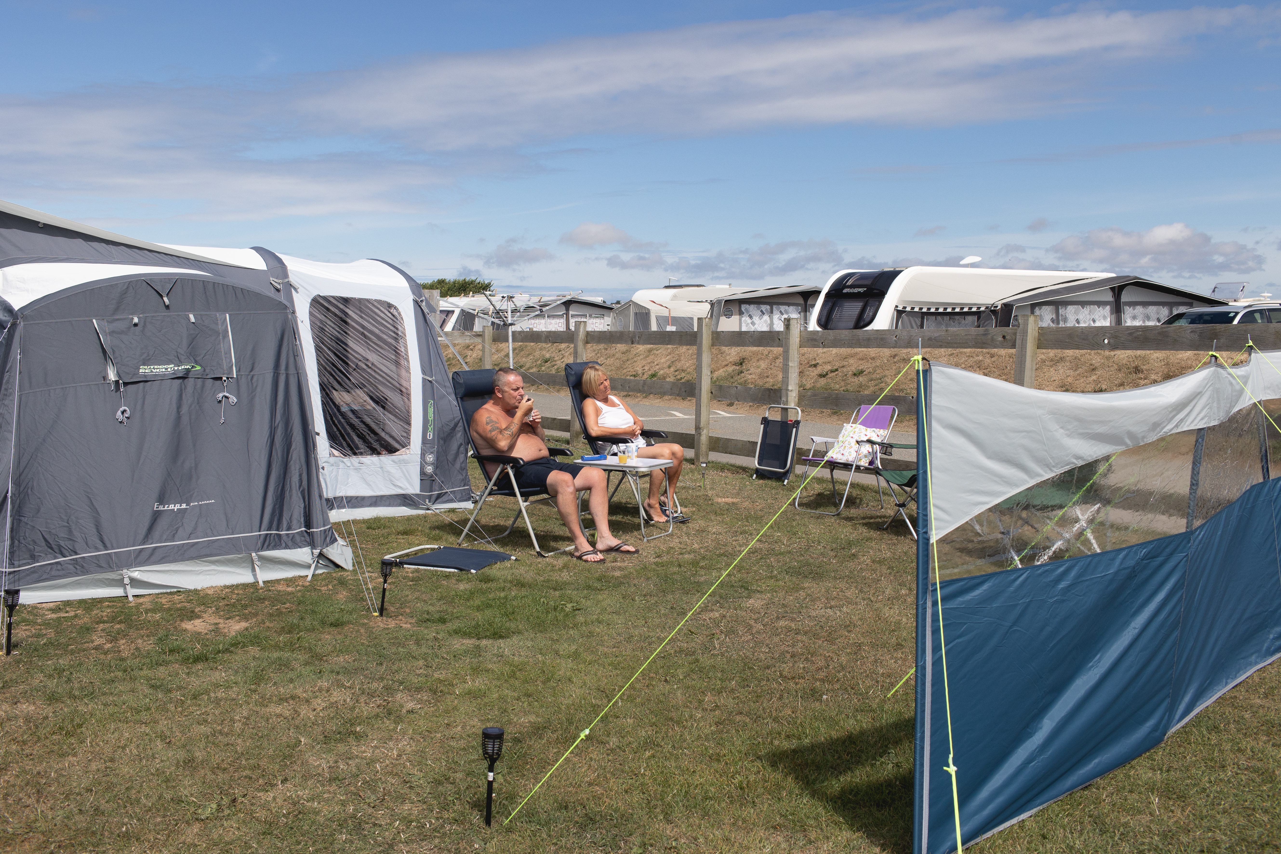 A photo of two people sitting on lawn chairs in front of a tent and surrounded by several tents and shades.