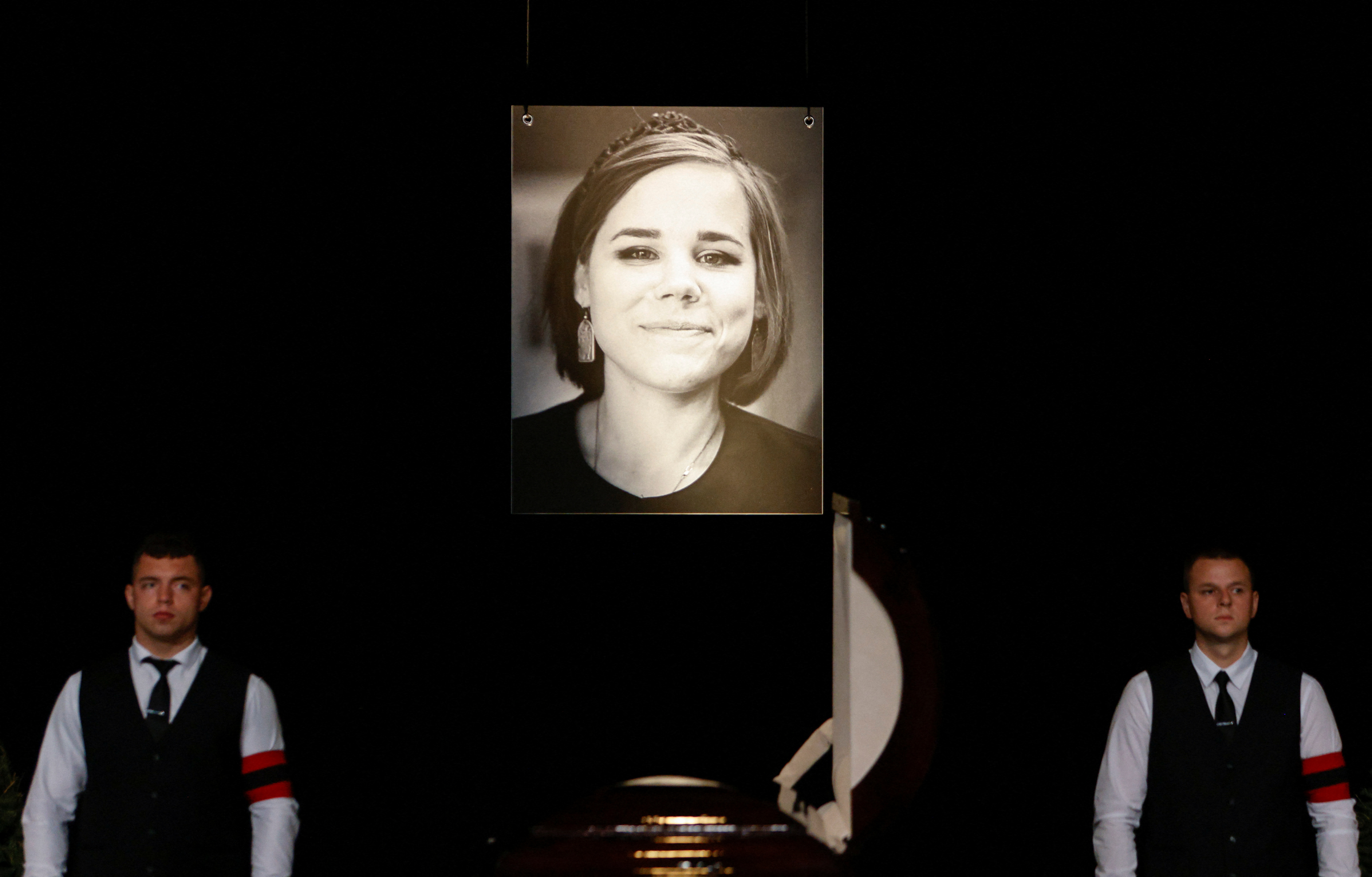 Memorial service for Darya Dugina with her coffin flanked by two men and a black and white portrait of her behind.
