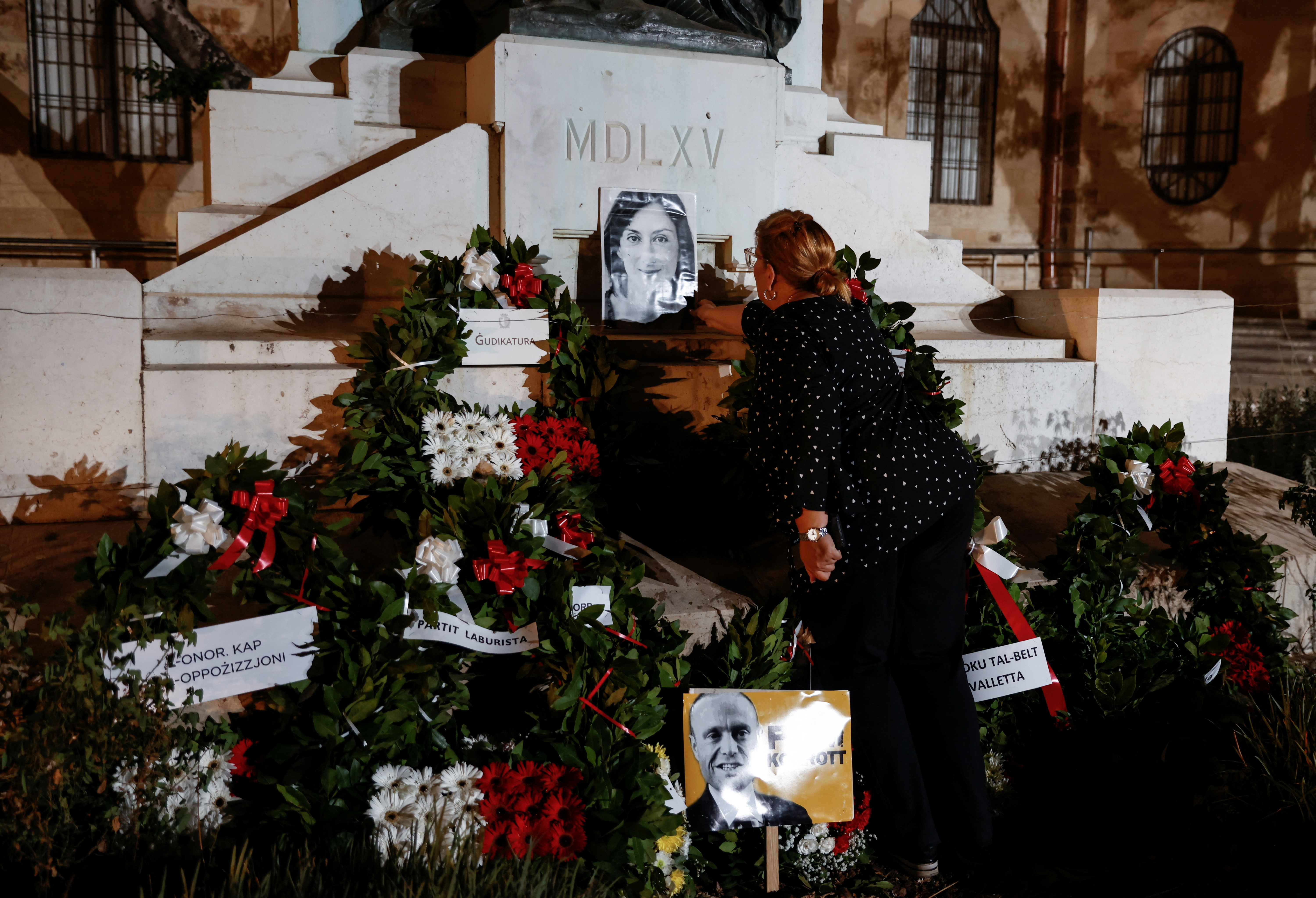 An activist places a portrait of assassinated anti-corruption journalist Daphne Caruana Galizia and a poster depicting former Prime Minister Joseph Muscat on the Great Siege Monument after an official wreath-laying ceremony, as people nearby protest against the authorities' lack of action over high-profile corruption cases first revealed by Caruana Galizia, in Valletta, Malta