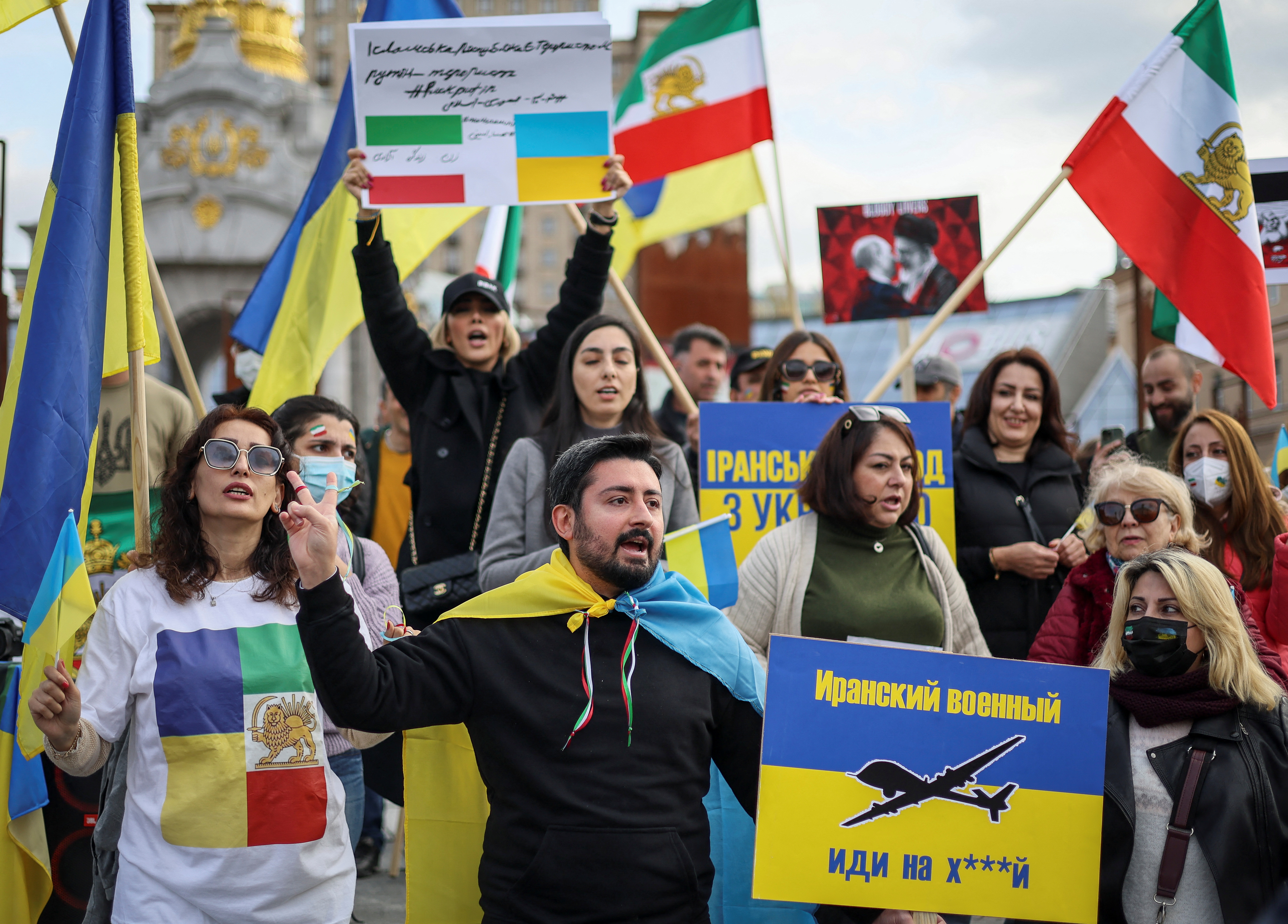 Protesters wearing Ukrainian colours and holding up Ukrainian and Irainian flags. One holds placard that says in Russia: 'Iranian serviceman, go f**k yourself'