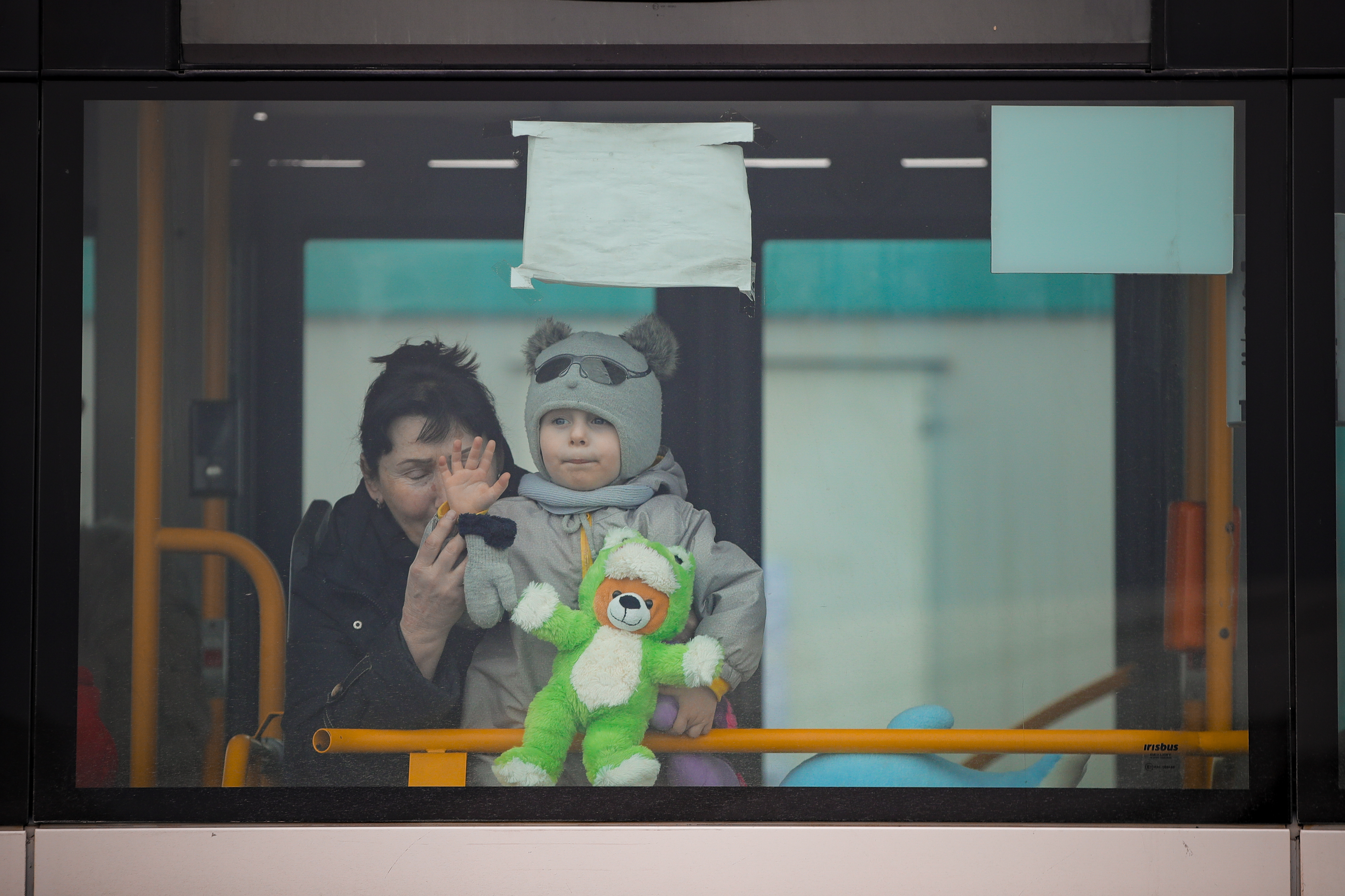 Boy is wearing hat with bear ears and holding green stuffed animal as he waves from the bus window, with his aunt helping to hold him up.