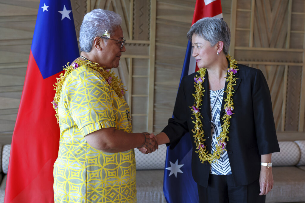 Australian Foreign Minister Penny Wong wearing a lei around her neck and in a black suit shakes hands with Samoa's Prime Minister Fiame Naomi Mata’afa who is wearing a yellow dress. There are flags in the background.