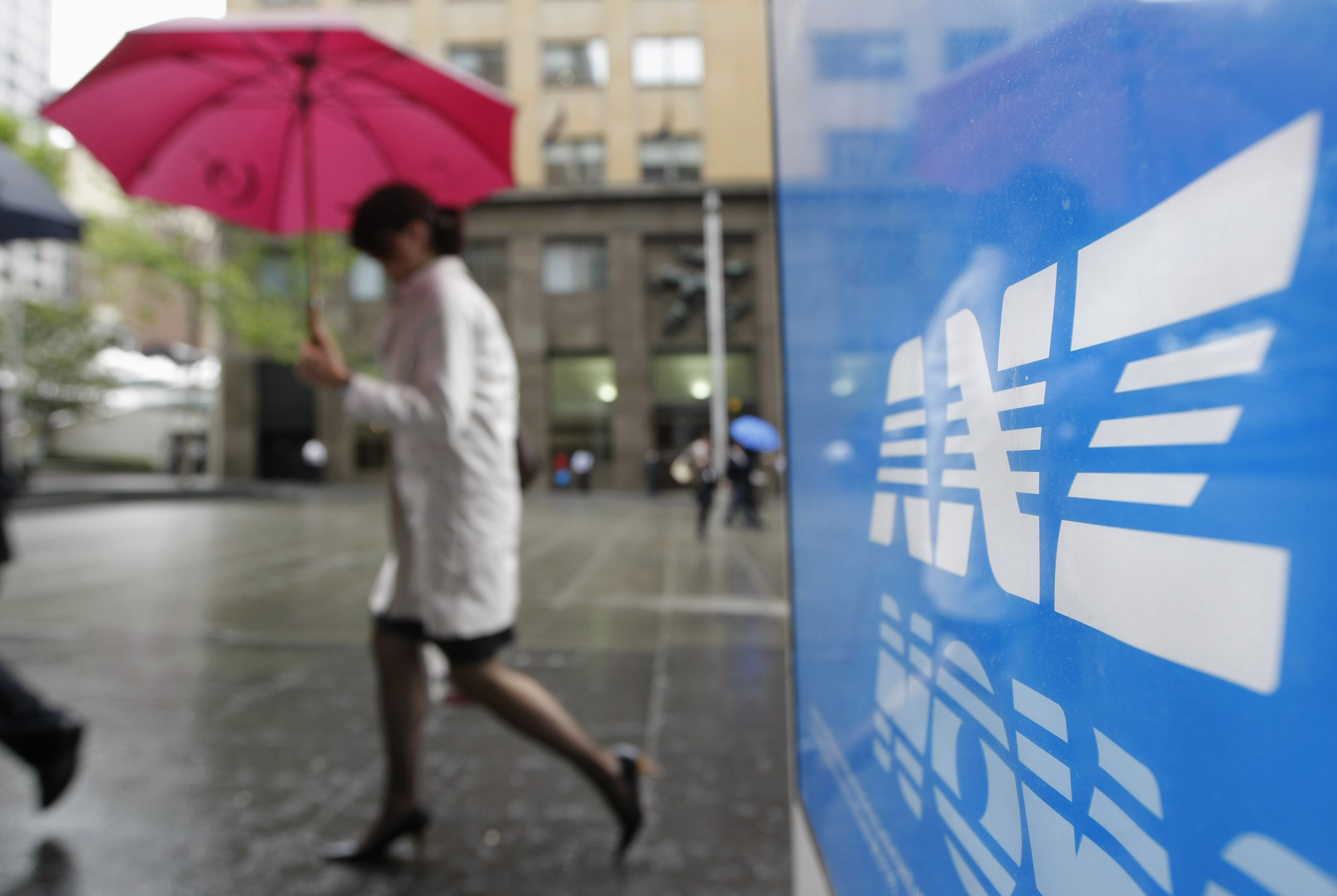 Close up of the blue ANZ sign with white letter and a blurred background of a rainy pavement, with woman walking past holding a red umbrella