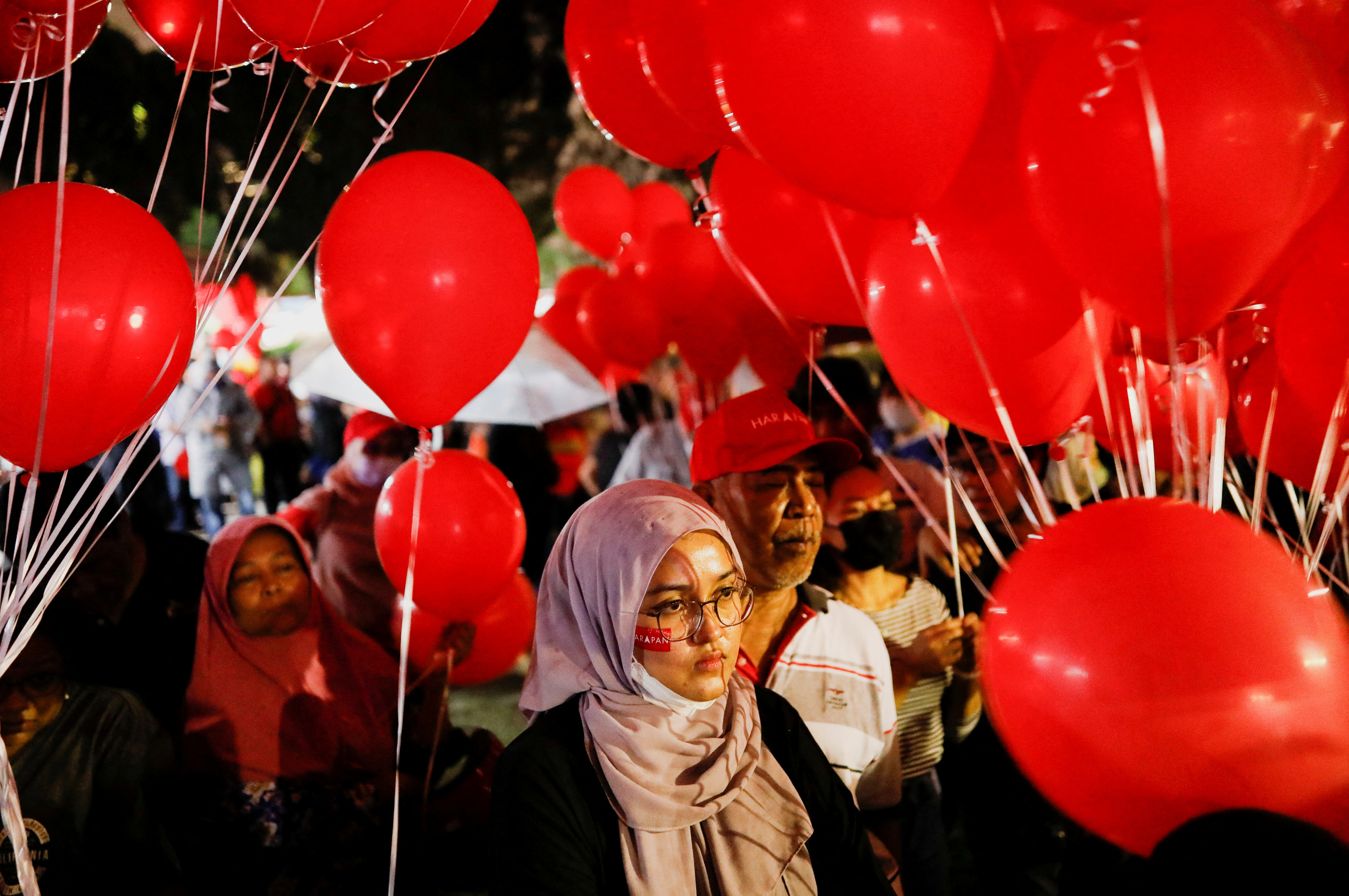 A young woman watching intently among a crowd of people with red balloons at a political rally for Pakatan Harapan